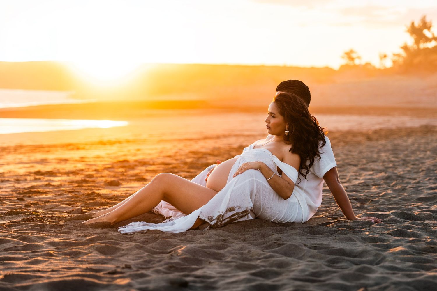 Pregnant woman in white dress sitting on sandy beach at golden sunset, cradling her baby bump.
