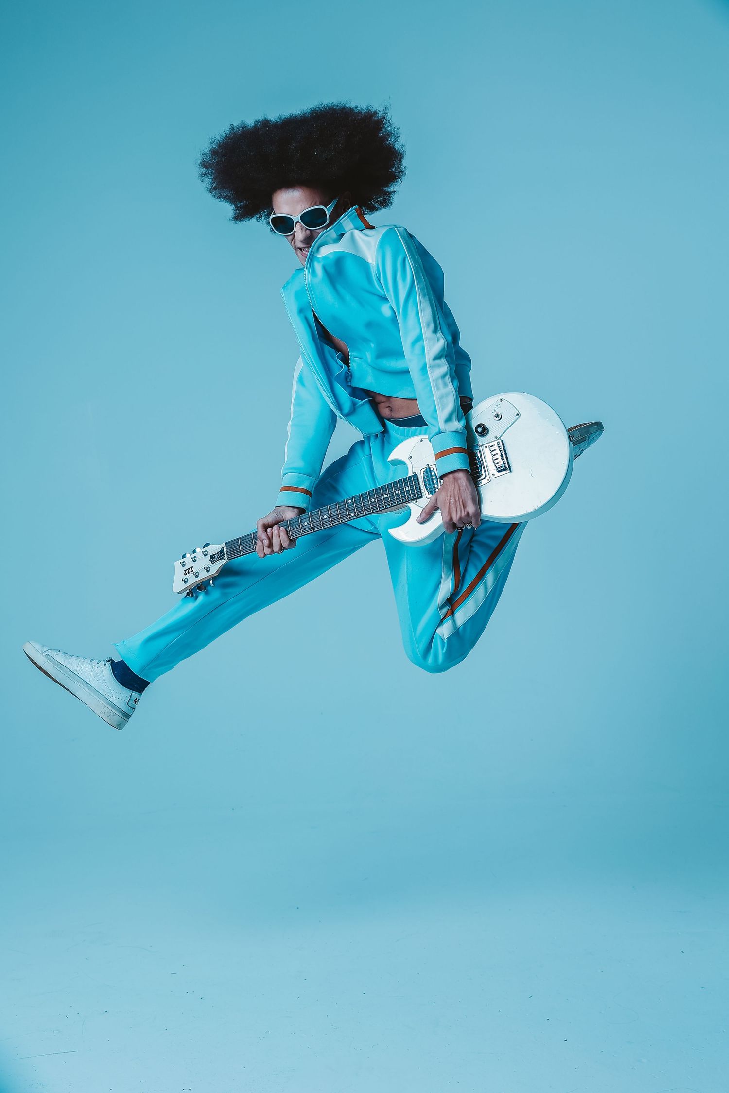 Performer in bright blue jumpsuit jumps with electric guitar against turquoise background.