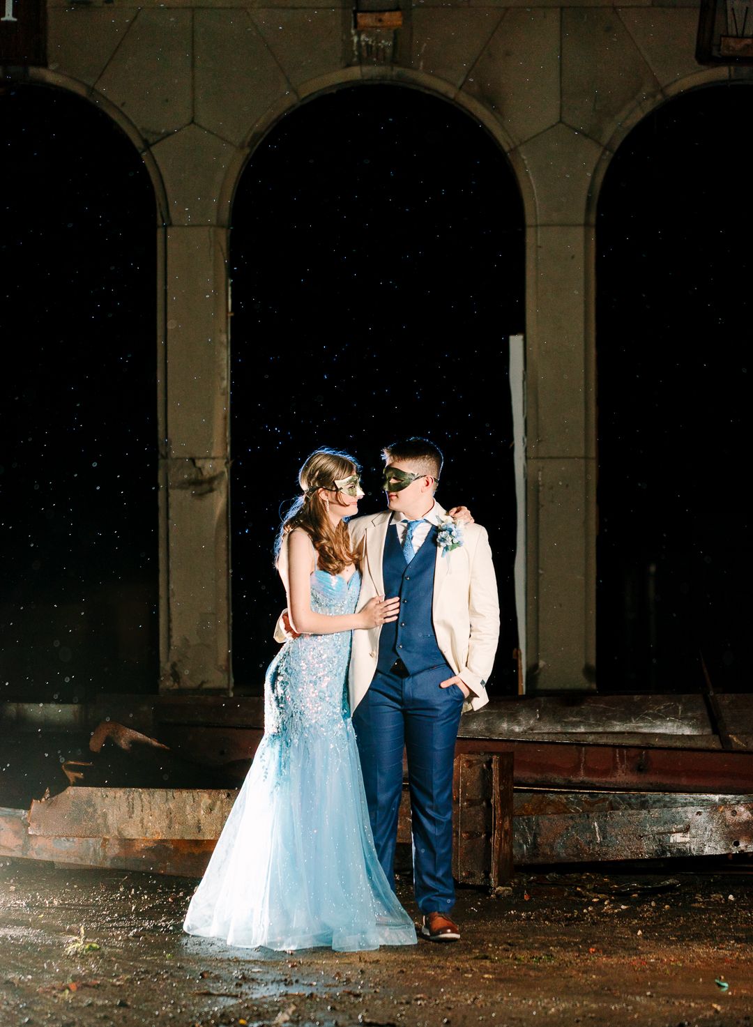 Elegant couple in wedding attire standing beneath stone archways at dusk in romantic architectural setting.