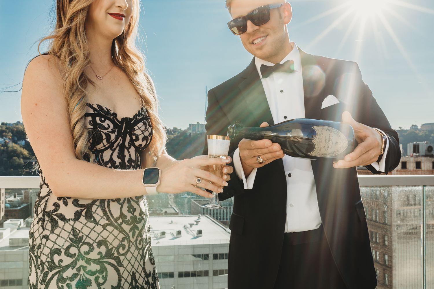 A couple in formal attire checks their watches on a sunny rooftop terrace.