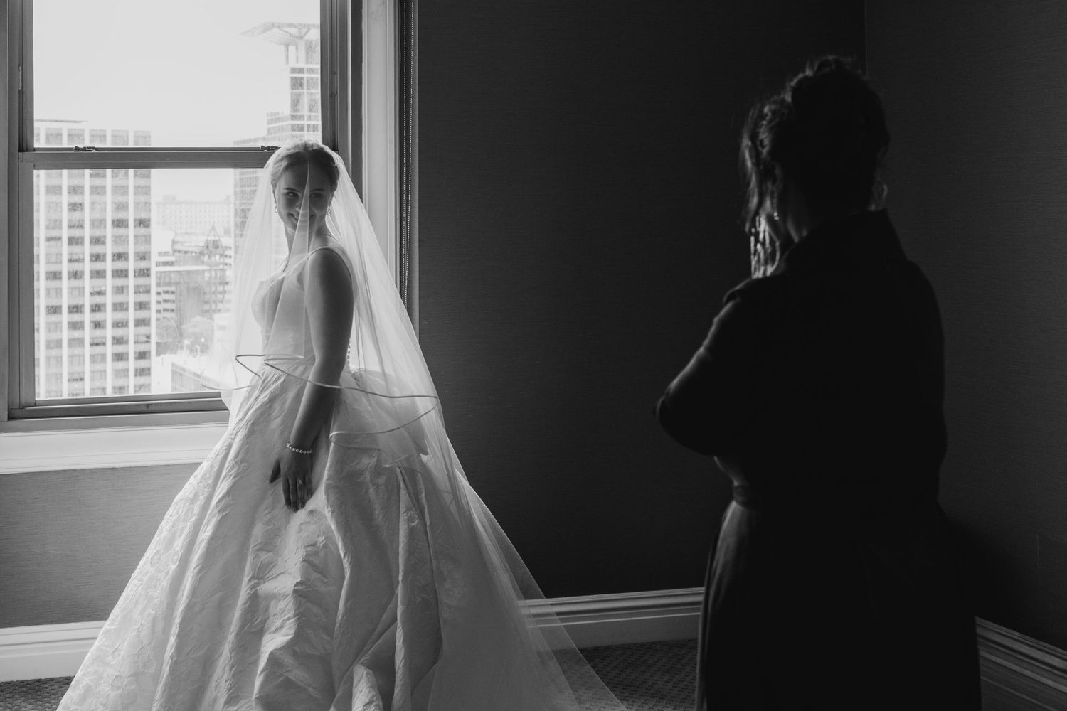 Black and white photo of a bride in a white gown and veil standing by a window with a silhouetted figure nearby.
