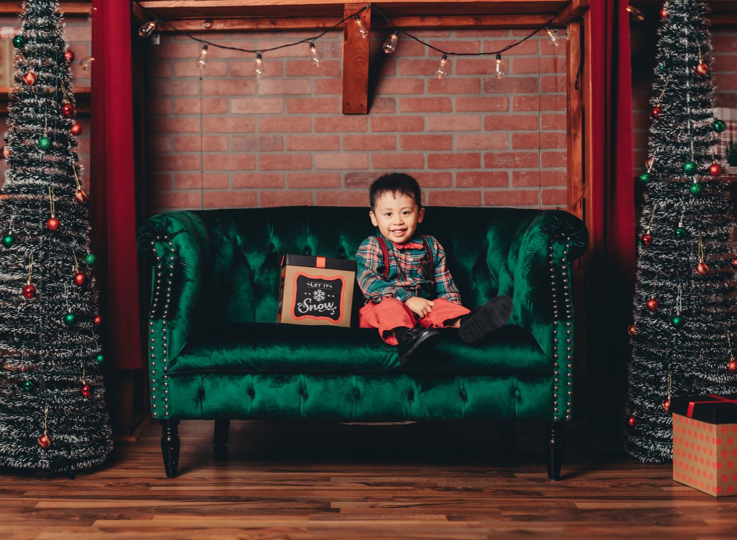 A festive holiday scene with emerald green velvet couches and sparkling Christmas trees against a brick wall.