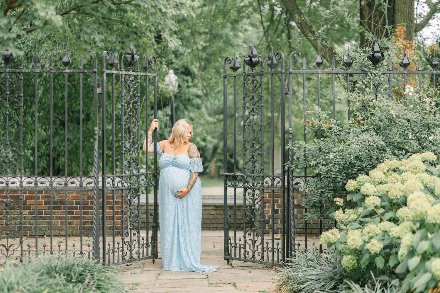Maternity photo of someone in a flowing blue dress standing between ornate garden gates.