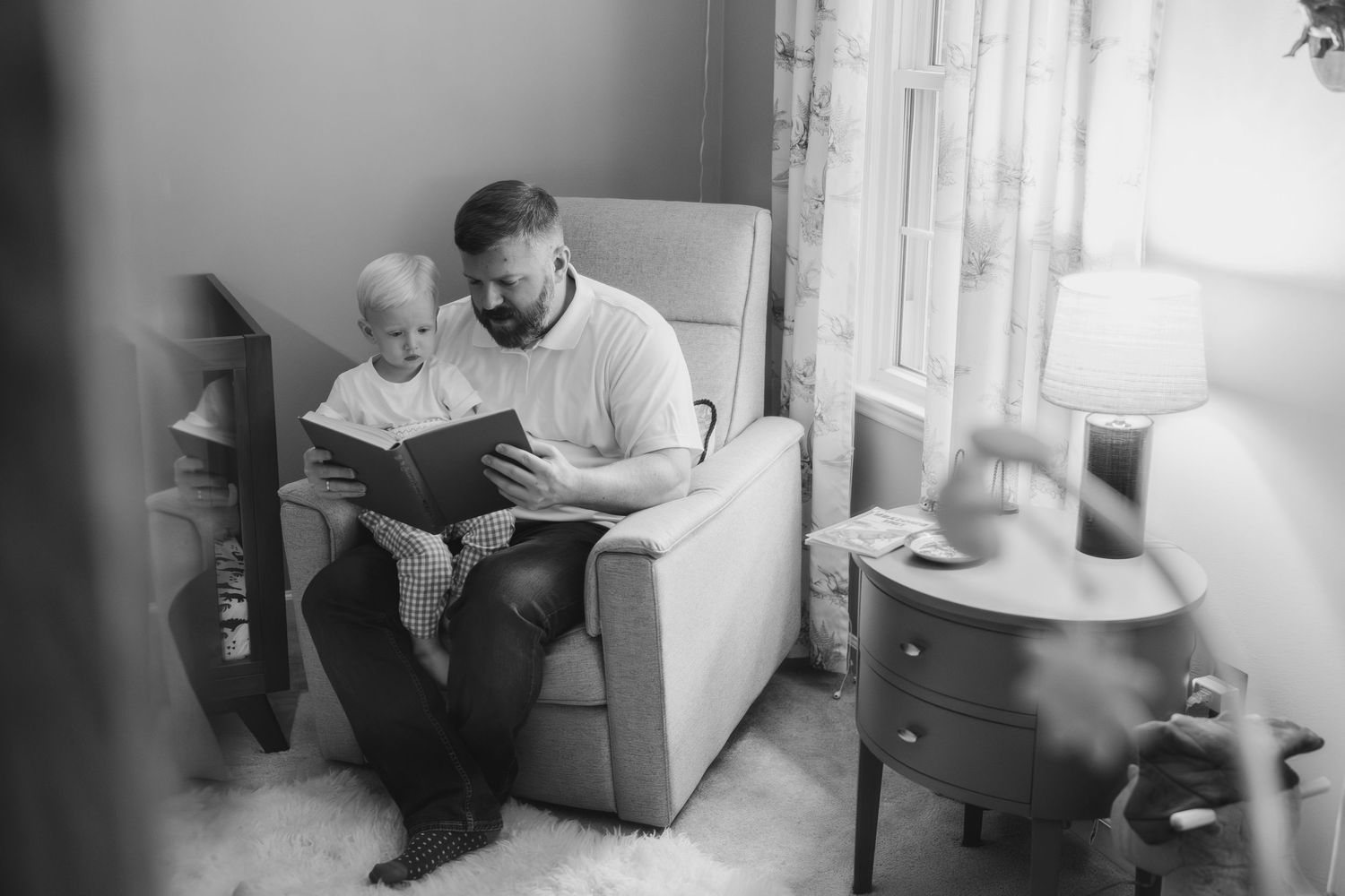 A person sits comfortably in a chair reading a book in a sunlit room with white curtains.
