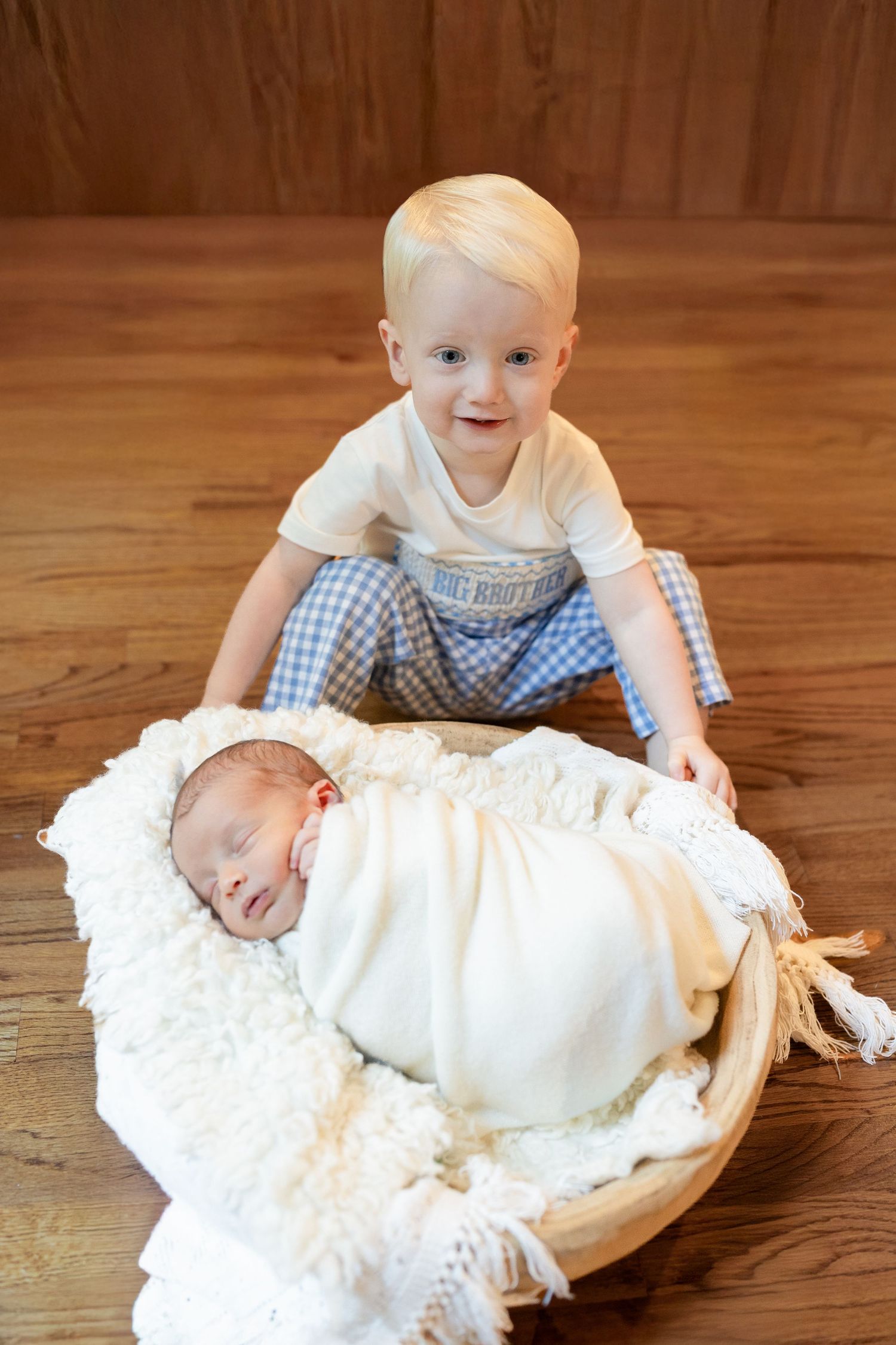 Two young children on hardwood floor with one looking at a sleeping baby wrapped in white blanket.