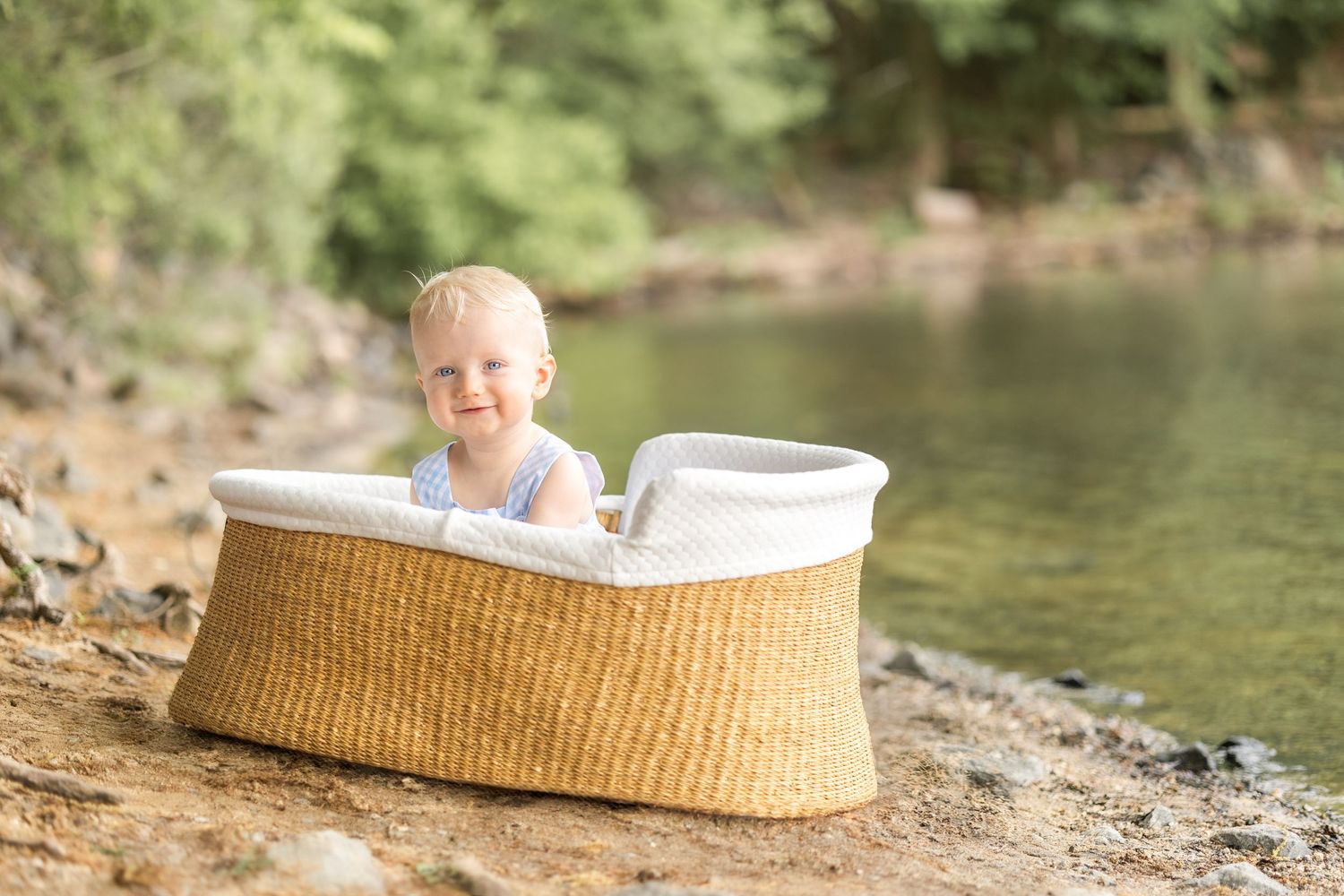 A baby lies in a woven basket outdoors near a peaceful stream with sunlight filtering through the trees.