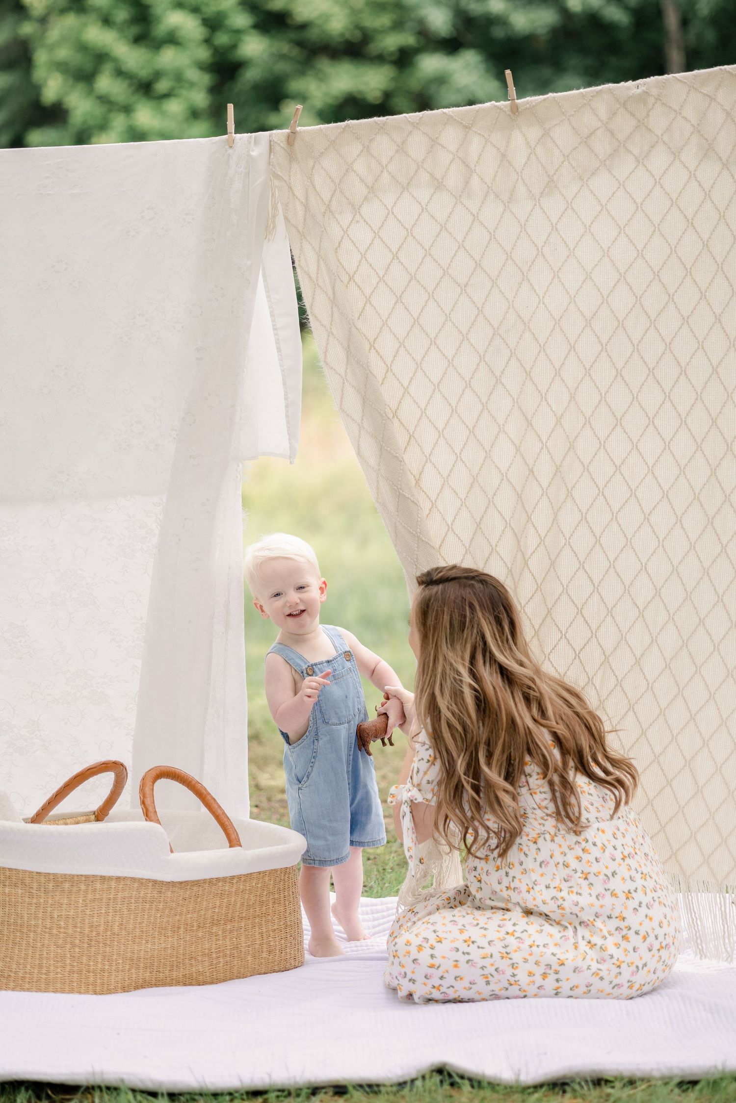 A baby in blue plays near white linens hanging to dry while someone watches from behind a patterned wall.