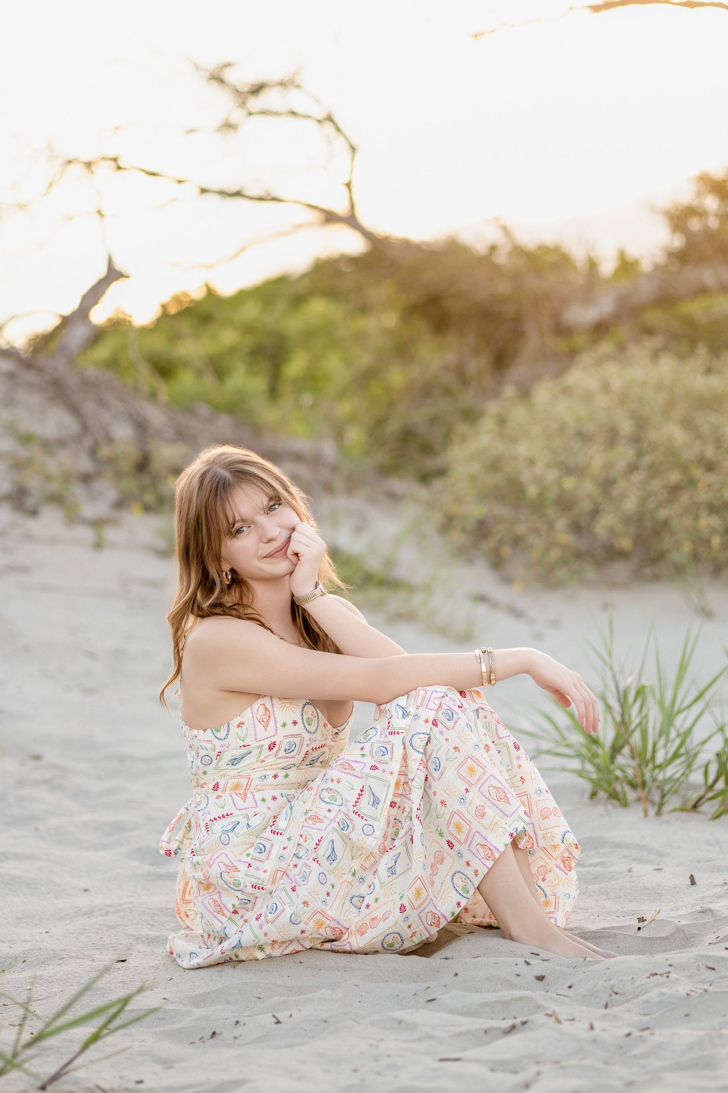 Young woman in floral dress poses playfully on sandy beach during sunset photo session.