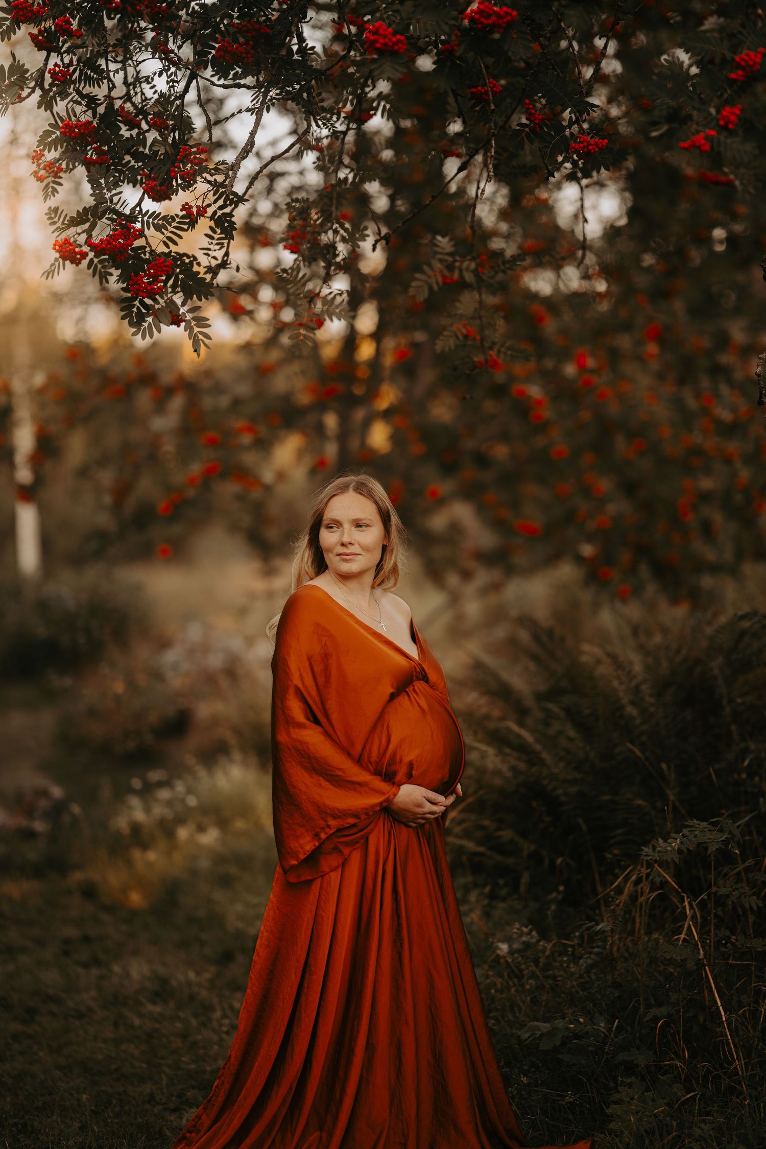 Expecting mother in stunning red gown poses beneath red berries in autumn setting.
