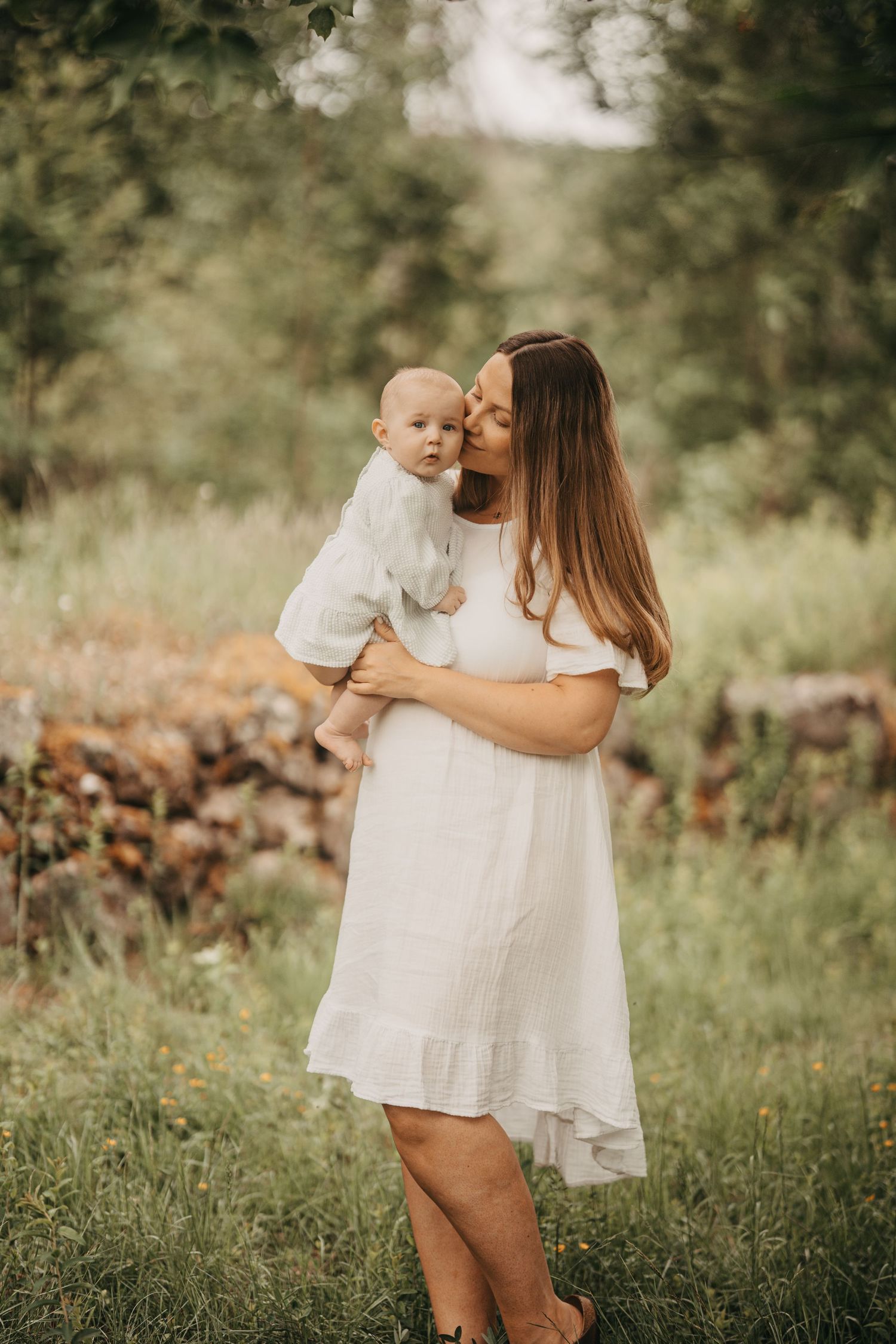 A mother in a white dress kisses her baby outdoors in a lush green garden setting.