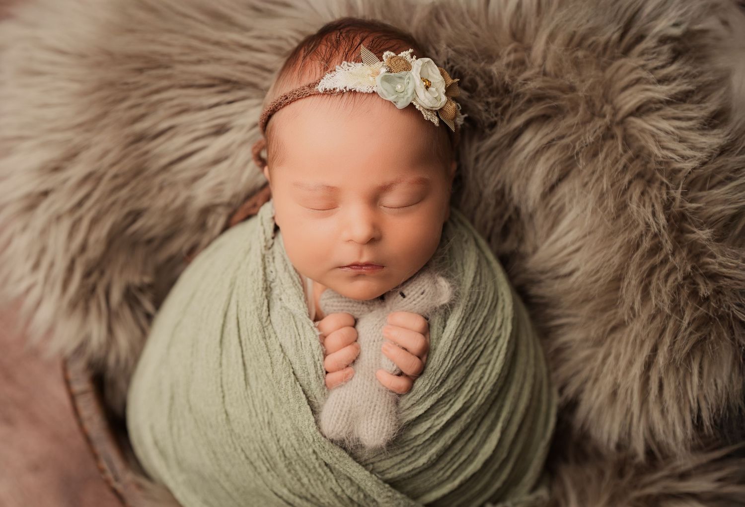 Sleeping newborn baby wrapped in green swaddle with floral headband holding tiny stuffed animal on fur blanket.