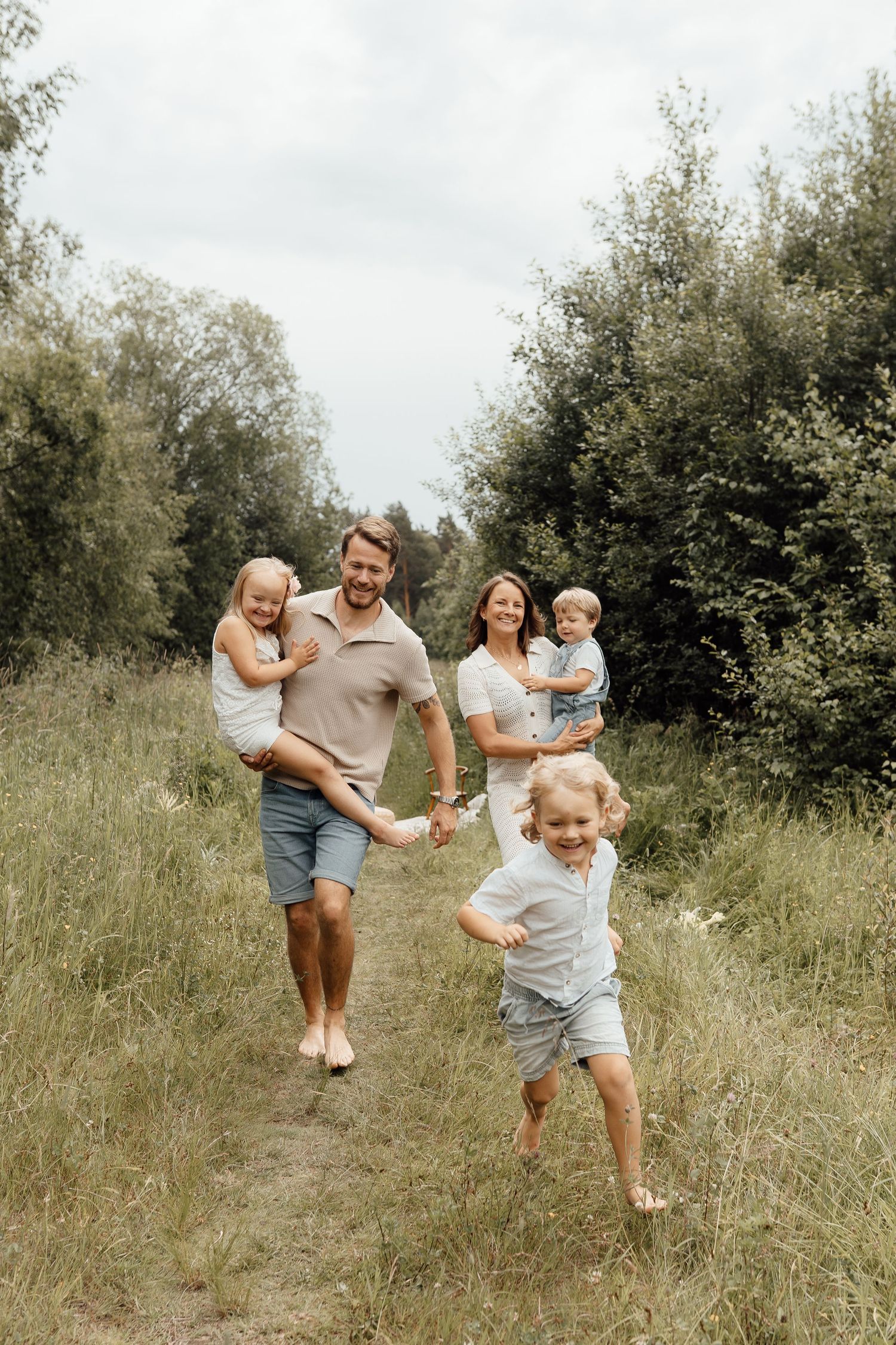 Happy family with two young children running and playing on a scenic grass path surrounded by lush green trees.