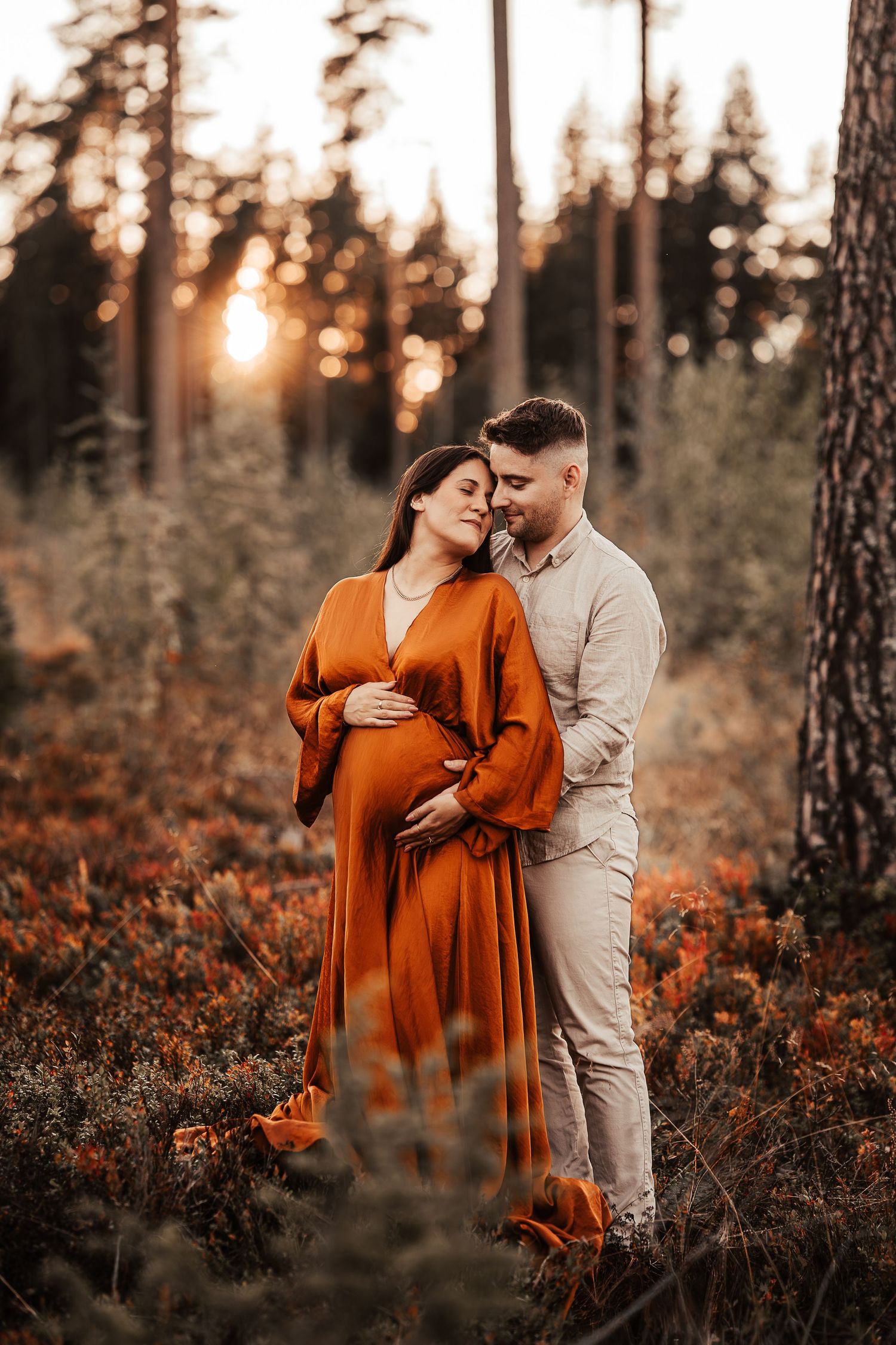A couple embraces in a forest at sunset with the woman wearing a flowing rust-orange dress against autumn foliage.