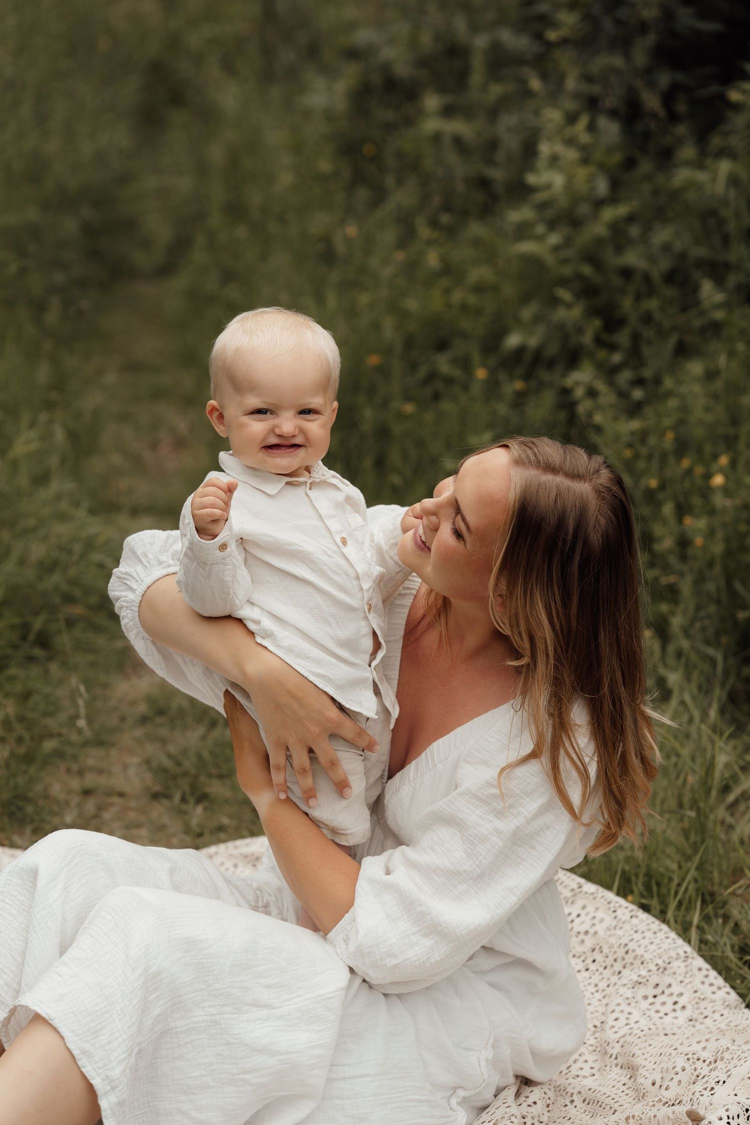 A smiling mother in a white dress holds her happy baby outdoors on a green meadow.