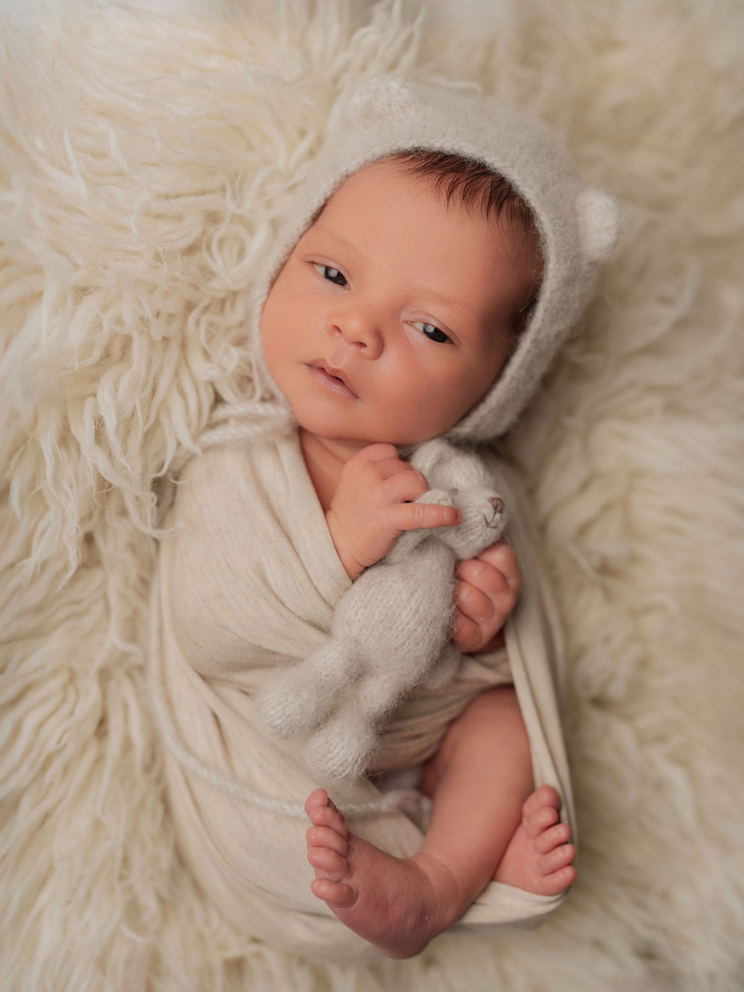 Newborn baby wrapped in cream knit swaddle wearing bear bonnet lying on fluffy white fur rug holding small toy.