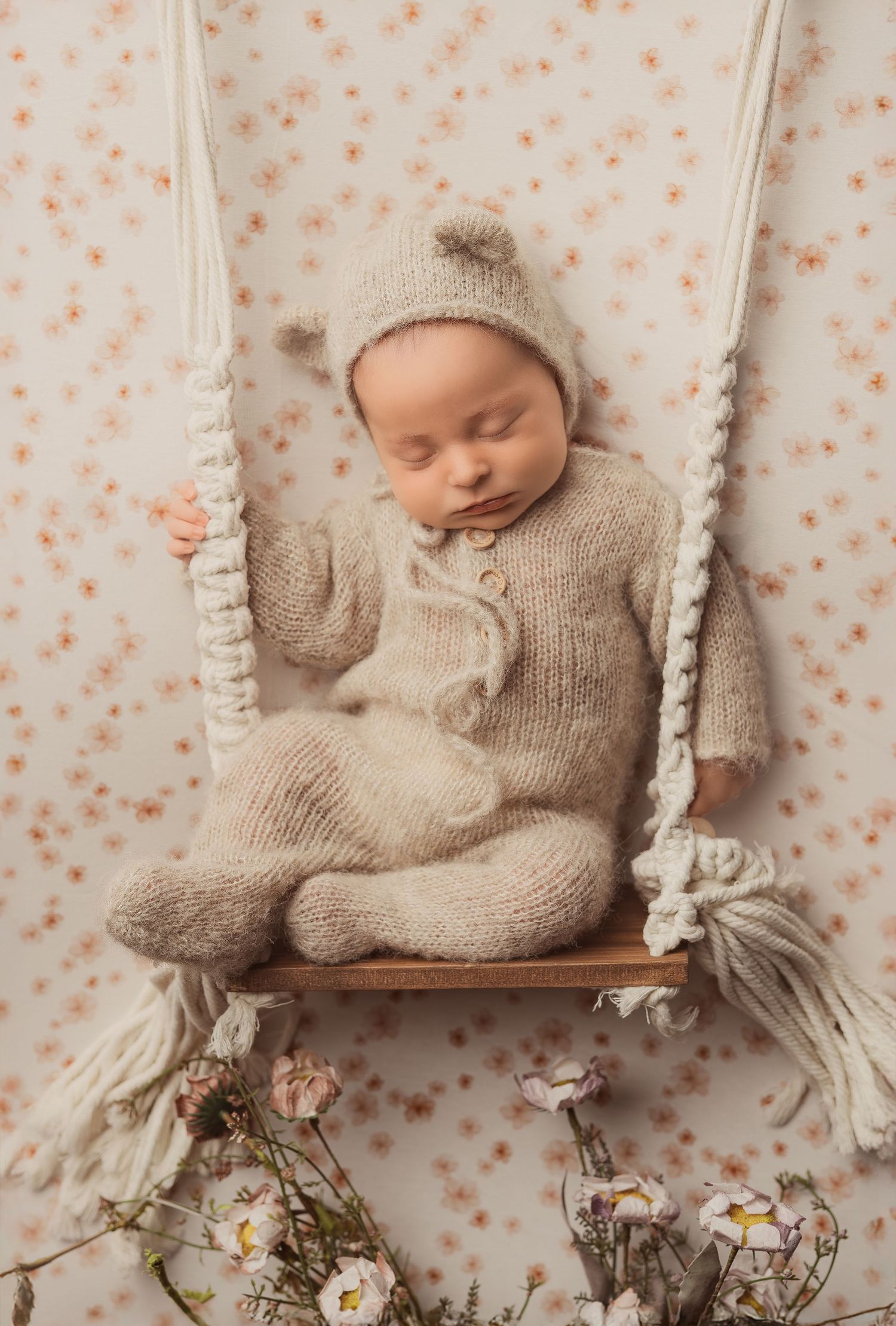 Sleeping newborn baby in beige knit outfit on macrame swing with floral backdrop and delicate flowers.