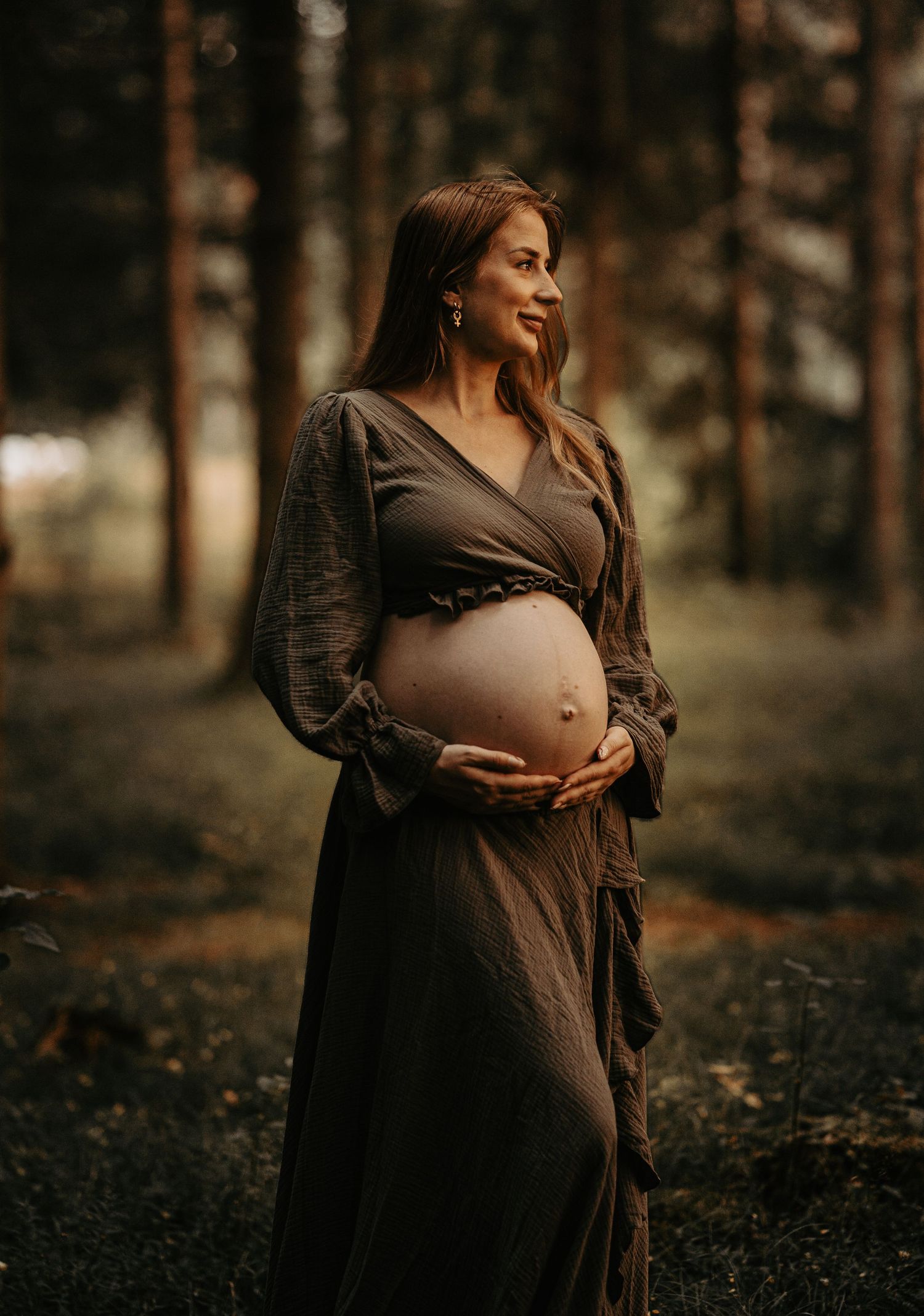Artistic maternity portrait in brown dress against forest backdrop.