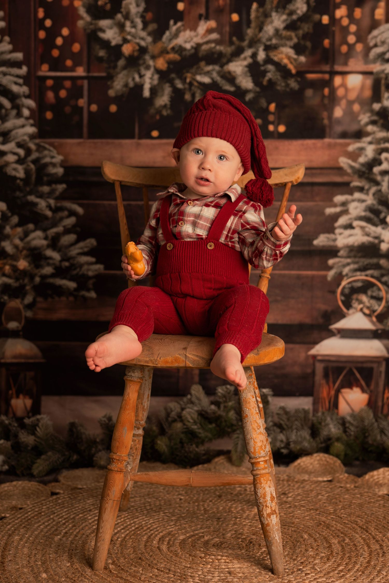 Baby wearing red knit outfit sits in rustic wooden chair against snowy winter backdrop with twinkling lights.