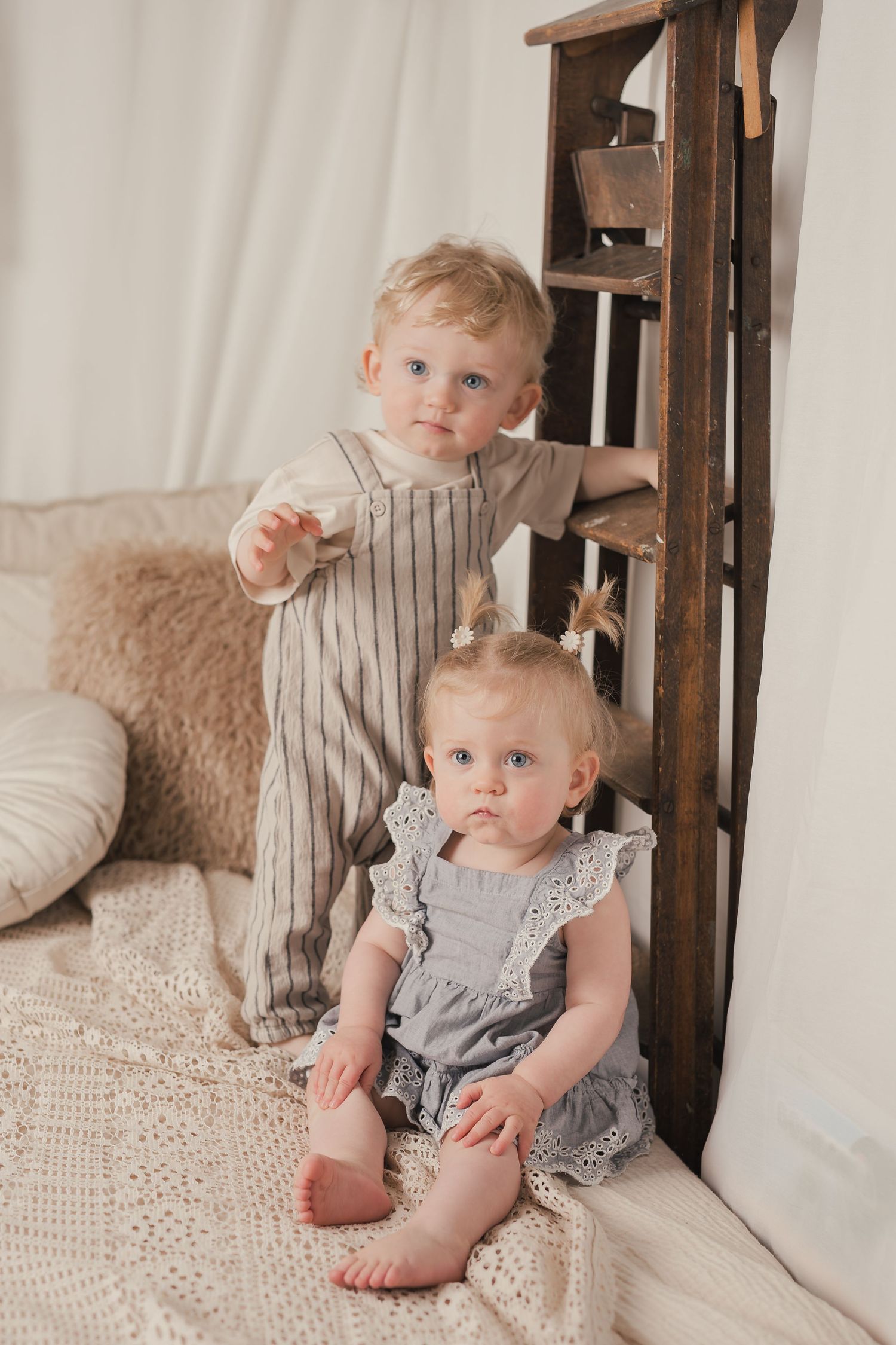 Two blonde toddlers in neutral linen outfits sit near a wooden ladder on a cozy white knit blanket.
