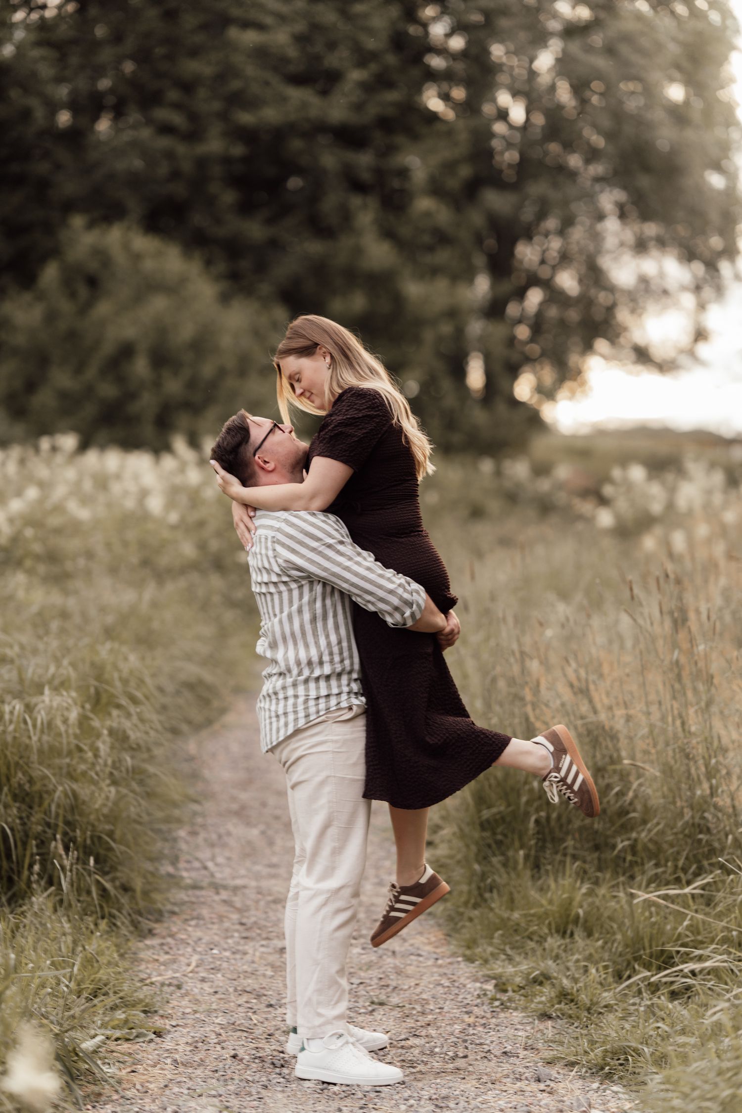 A man lifts a smiling woman on a scenic path surrounded by tall grass and trees during a romantic outdoor couples photoshoot.