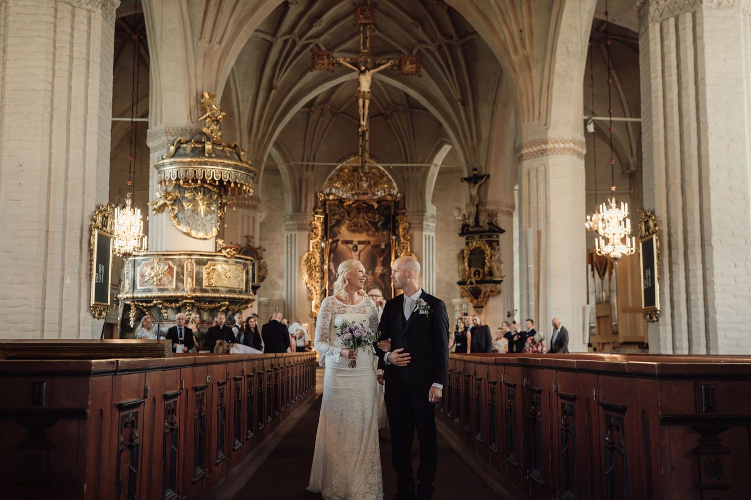 Bride and groom walking down the aisle of an ornate historic church with golden decor during wedding ceremony.