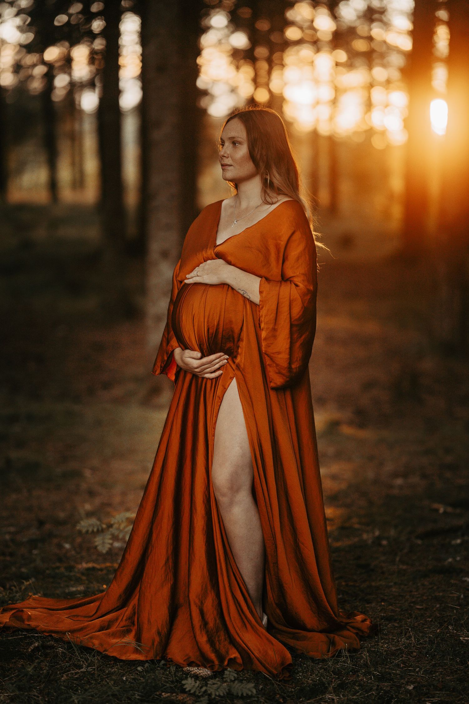 Dramatic maternity portrait in flowing orange dress with sunset forest backdrop.