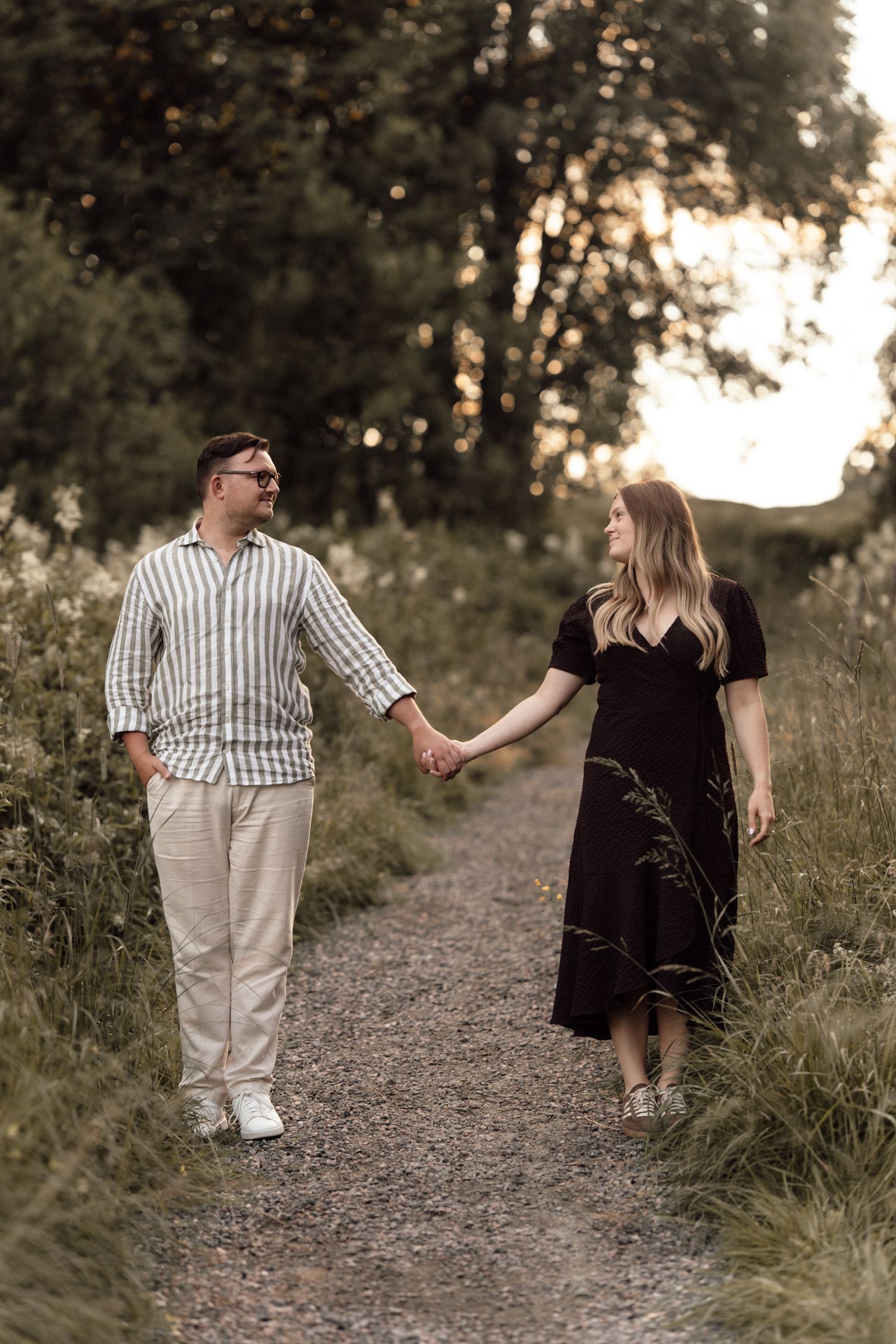 Couple holding hands walking on a gravel path surrounded by lush greenery at golden hour sunset.