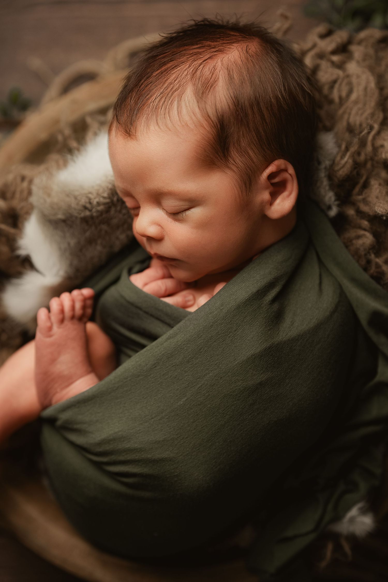 Peaceful newborn baby swaddled in a green wrap sleeping in a rustic wooden bowl with soft fur props.