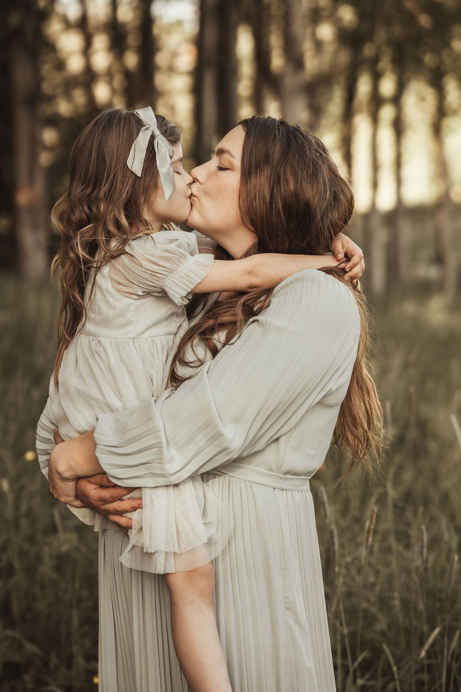 A mother holds and kisses her young daughter outdoors in a forest, both wearing matching white dresses.