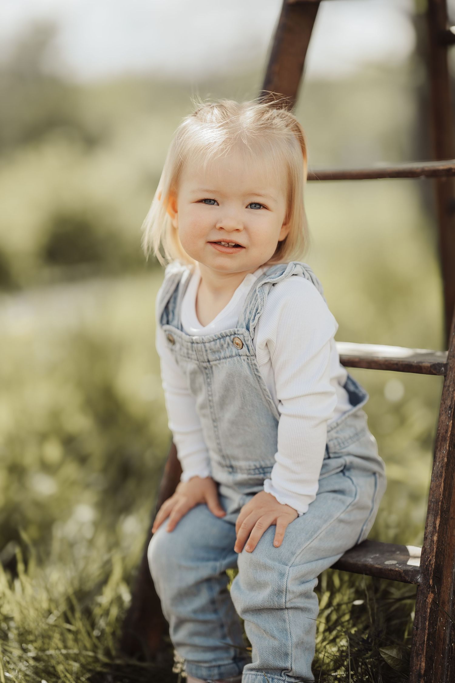 Smiling toddler girl in denim overalls and white shirt sitting on a wooden ladder in a green outdoor setting.