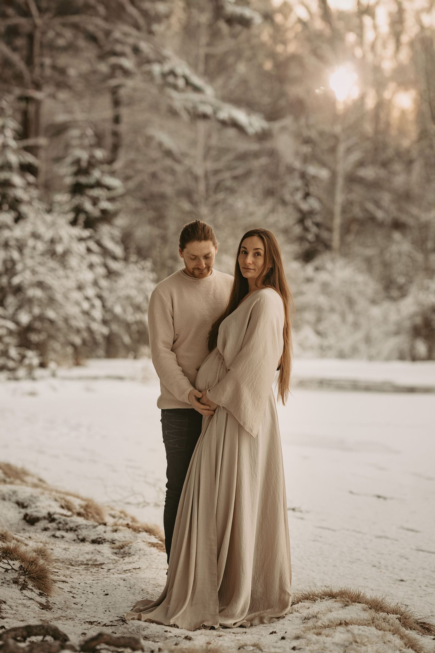 Winter maternity session with couple in neutral colors against snowy forest backdrop.