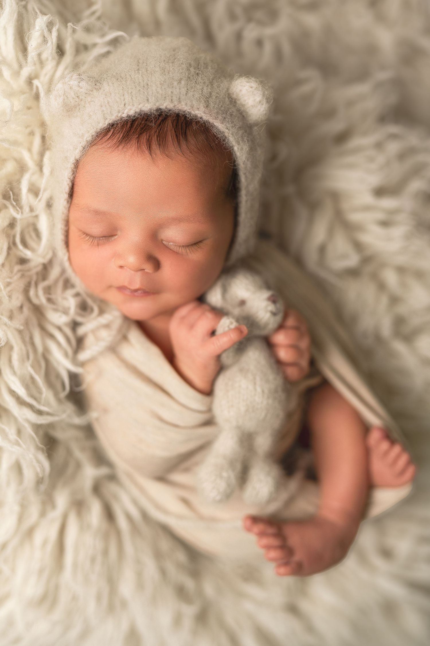 Sleeping newborn baby in cream outfit and bear bonnet cuddles a tiny teddy bear on a fluffy white fur blanket.