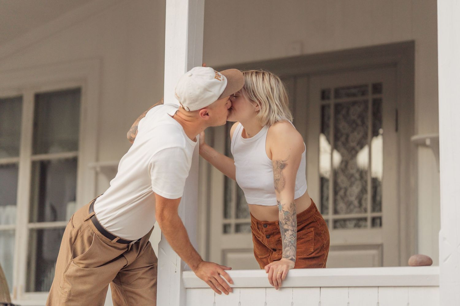 Couple sharing a romantic kiss on a white porch railing outside a house.