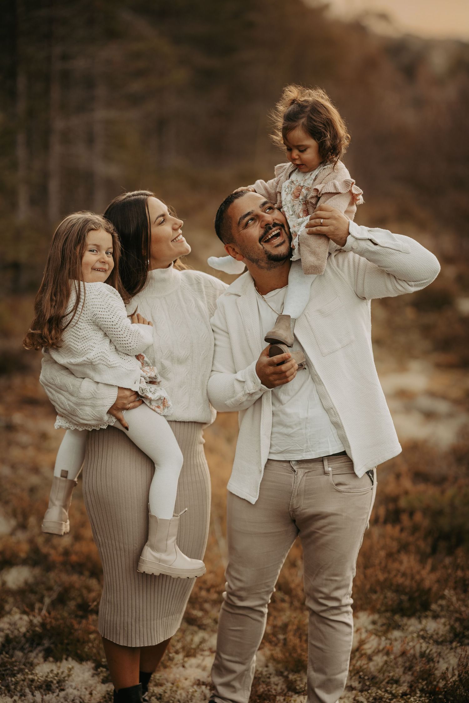 A happy family of four embraces outdoors during golden hour in coordinated white and neutral outfits.
