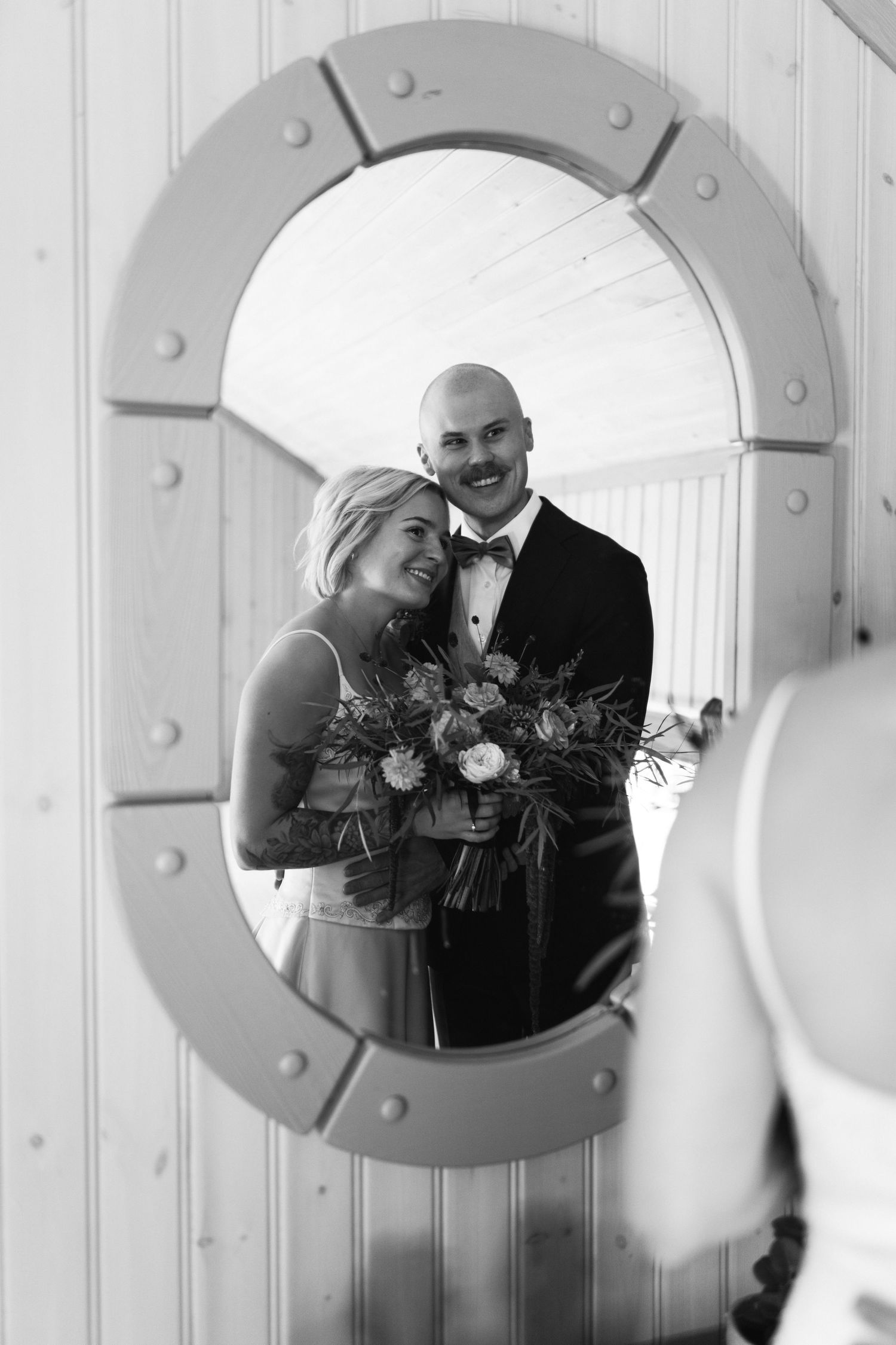 Black and white wedding photo of smiling bride with bouquet and groom in tuxedo reflected in ornate mirror.