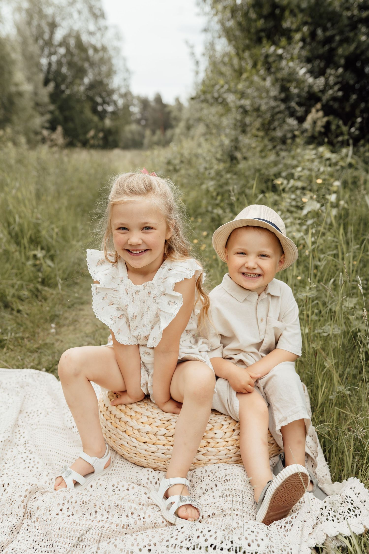 Two smiling children sitting on a wicker basket on a blanket in a sunny green meadow, dressed in neutral summer outfits.