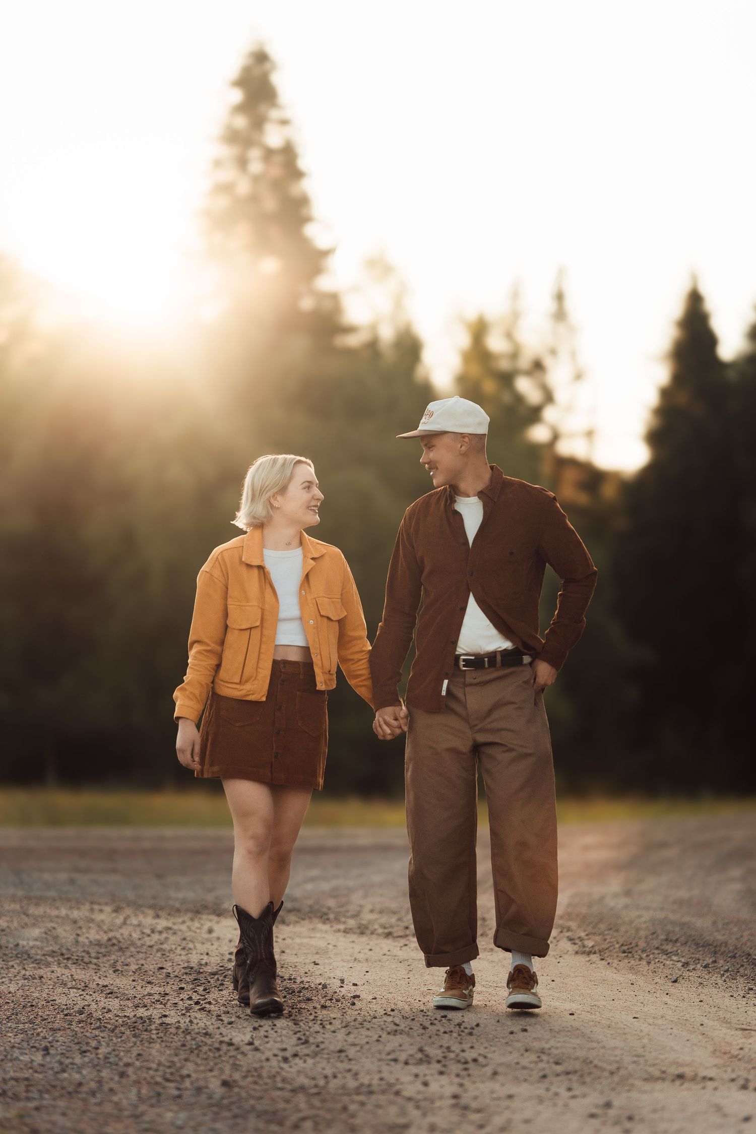 Couple holding hands walking on gravel path at golden hour, woman in orange jacket and brown skirt, man in brown outfit.