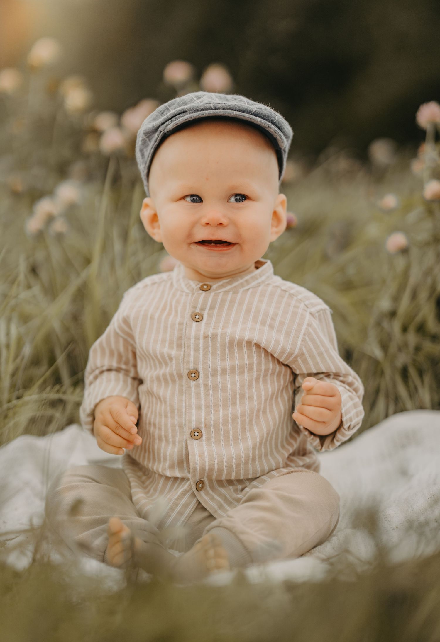 Smiling baby boy in striped outfit and flat cap sitting in a sunny flower meadow.