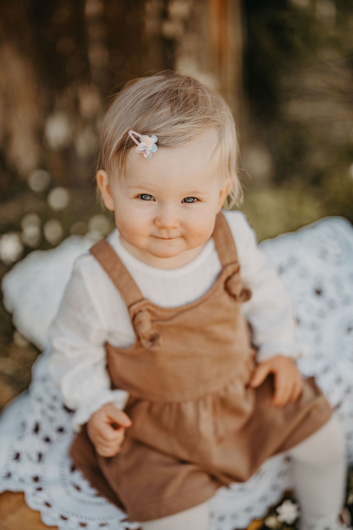 Adorable baby girl in brown overalls and white shirt with hair clip smiling outdoors on lace blanket.