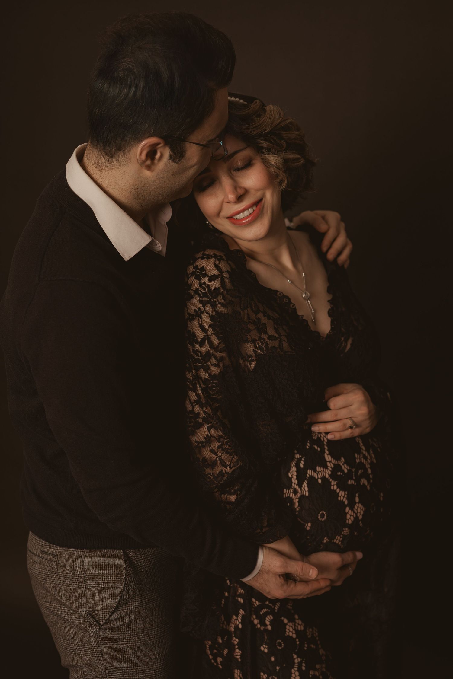Intimate maternity portrait with couple in formal black attire against dark background.