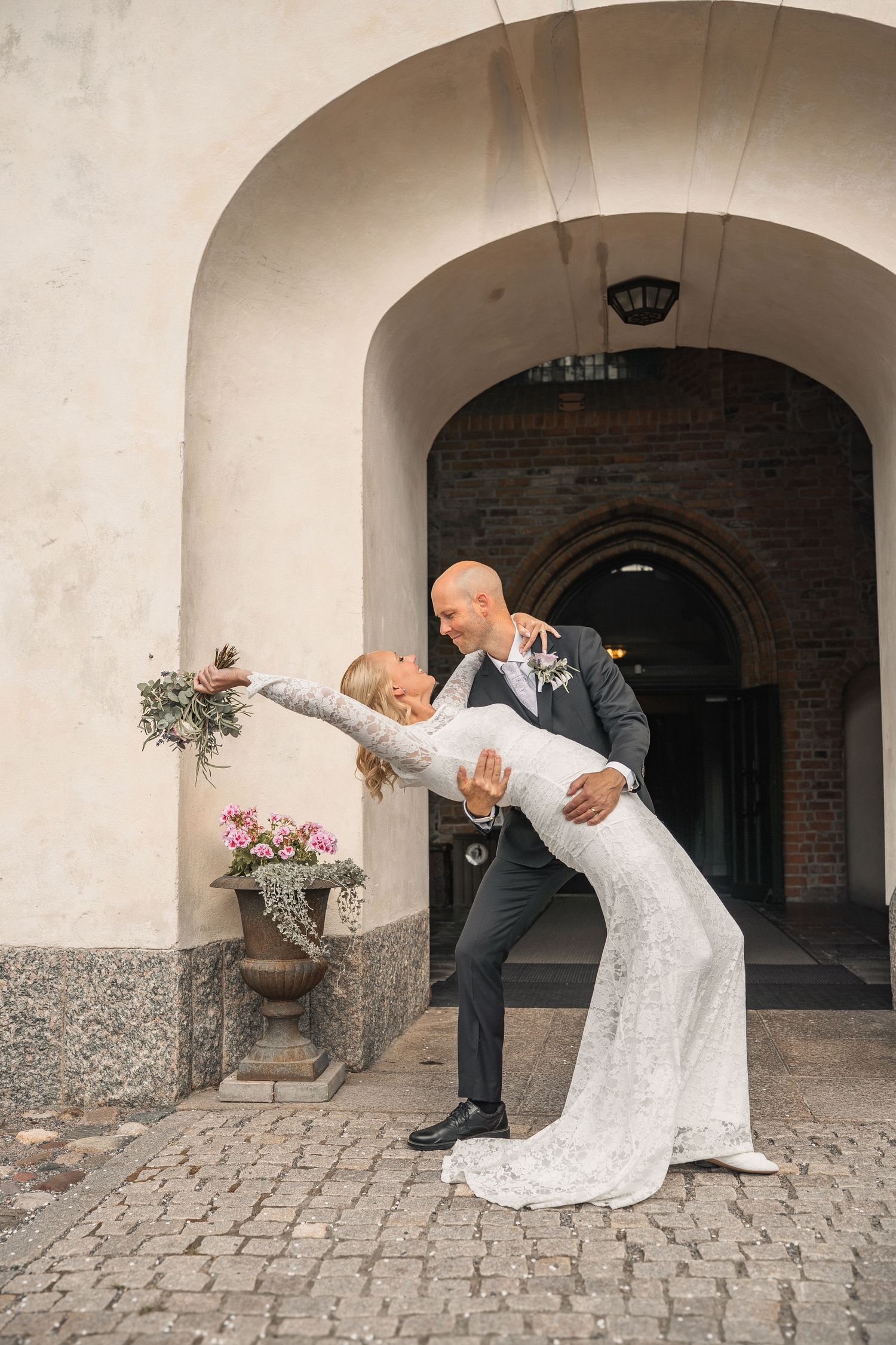 A groom dips his bride in a white lace gown under an archway on cobblestones, as she holds her bouquet out.