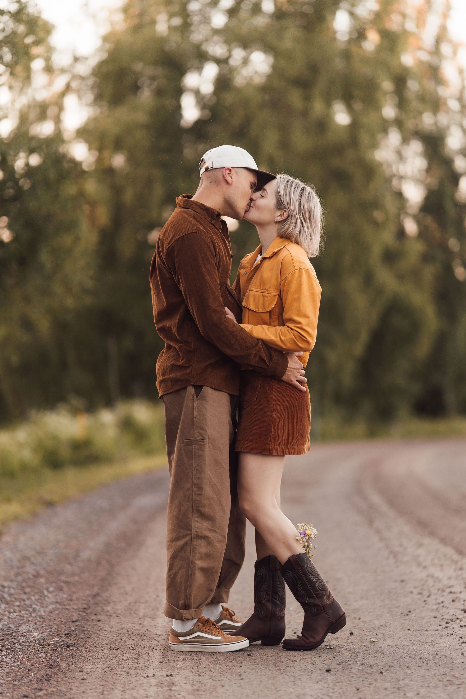 Couple sharing a romantic kiss on a rural dirt road surrounded by lush green trees in warm autumn tones.