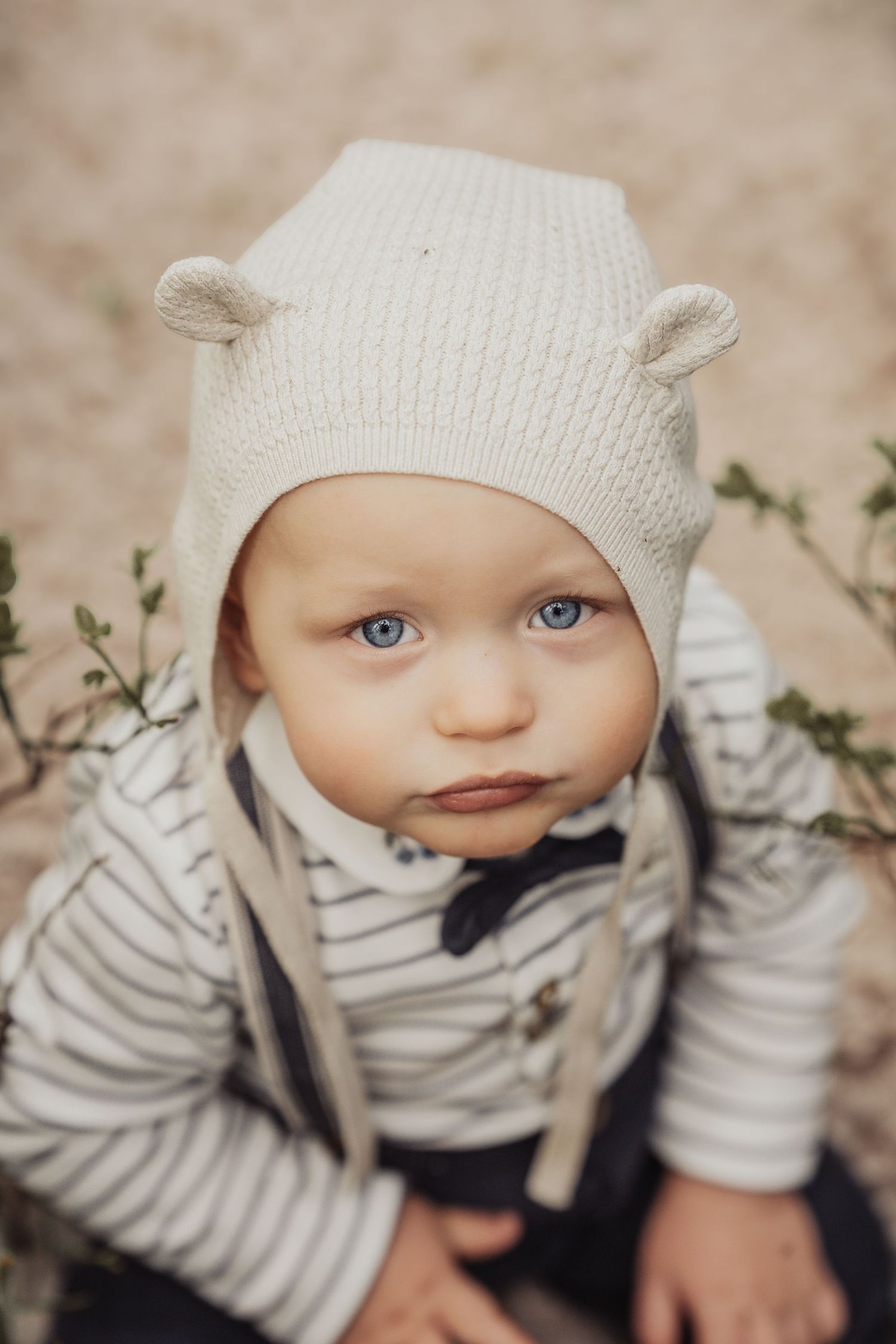 Adorable baby with blue eyes wearing a cream knit bear ear hat and striped shirt outdoors.