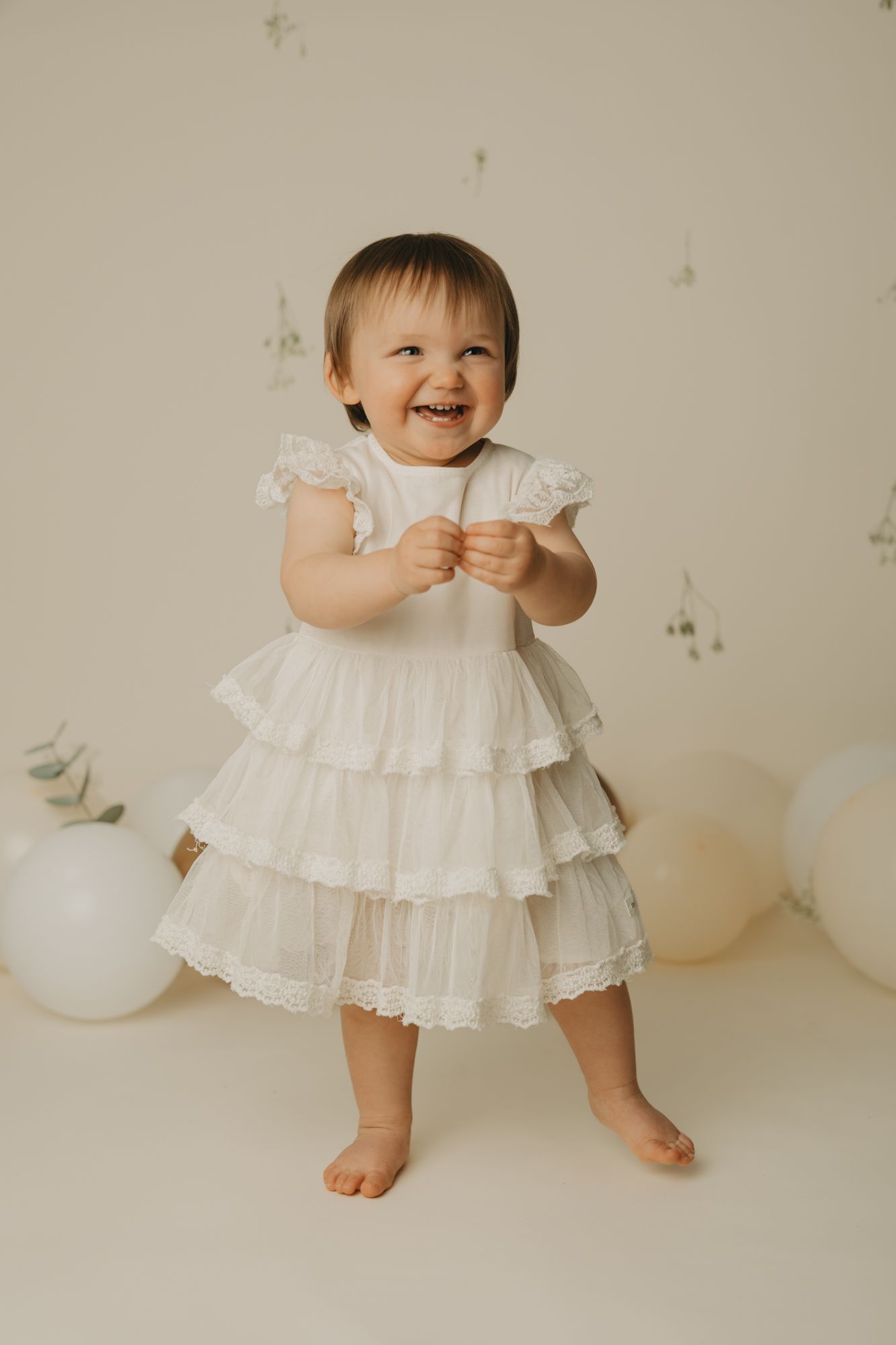 Baby wearing white ruffled dress stands barefoot against neutral background with white balloons.