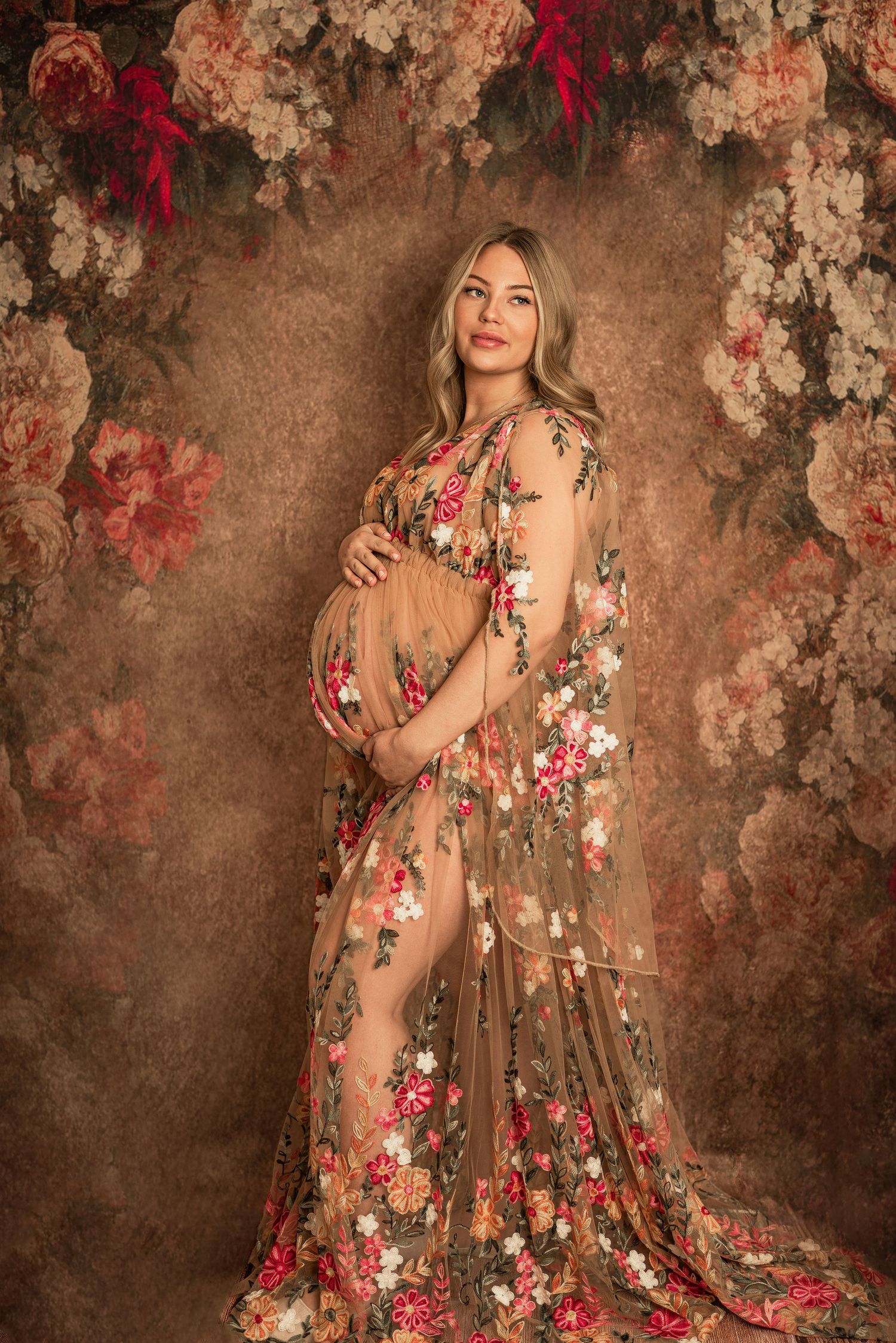 A maternity portrait with a sheer floral gown against a rustic brown backdrop.