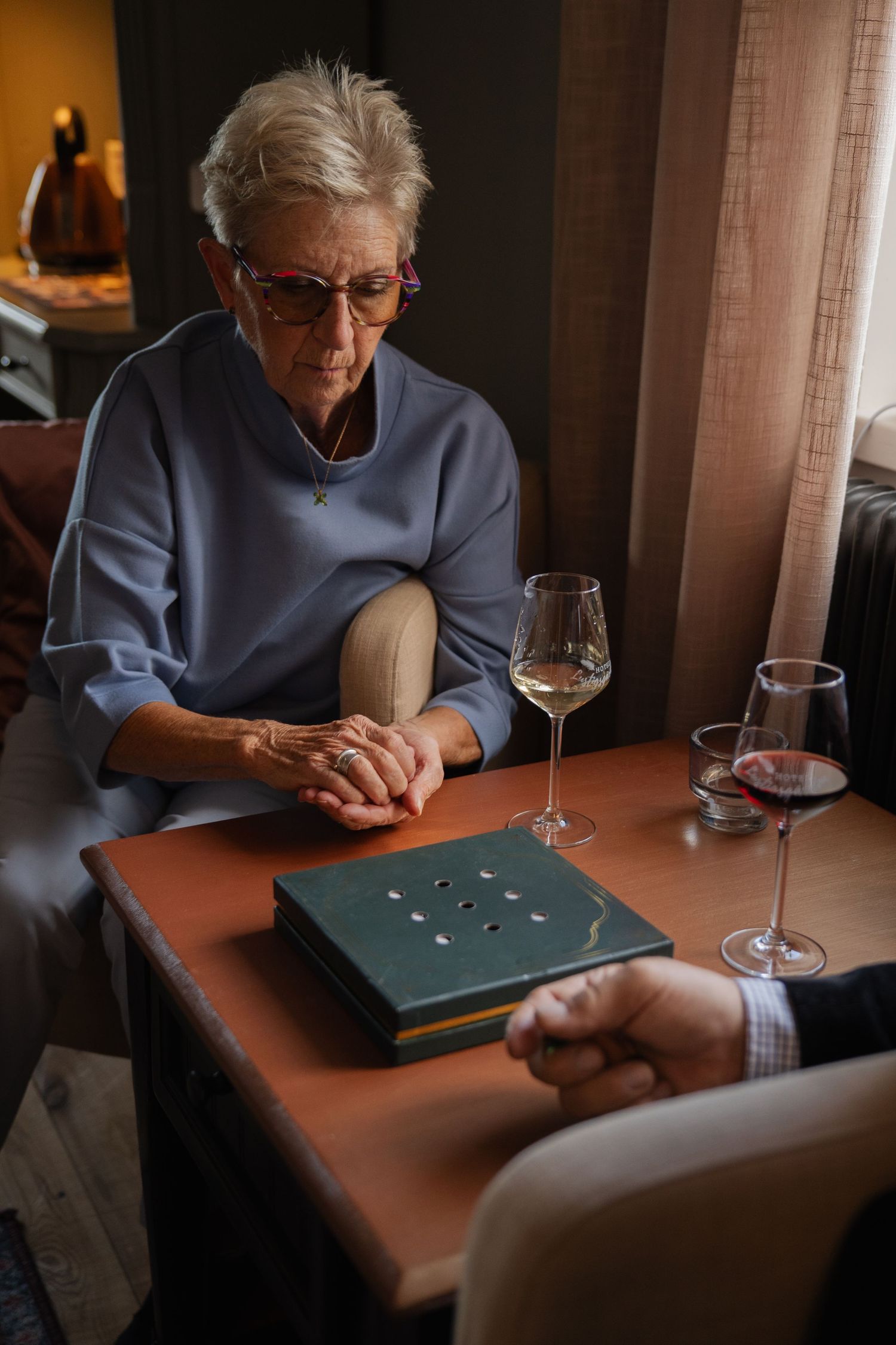 Person wearing gray sweater examines small objects on a dark table with a wine glass nearby in dim lighting.