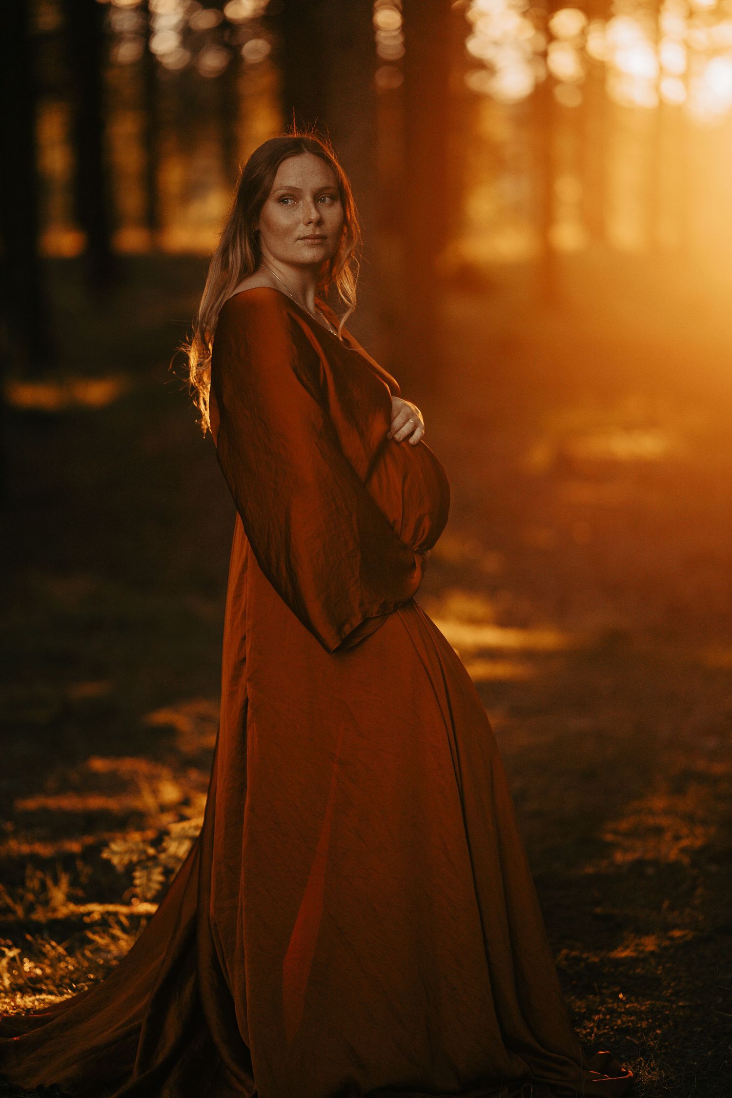 Sunset maternity portrait in rust-colored gown among forest trees.