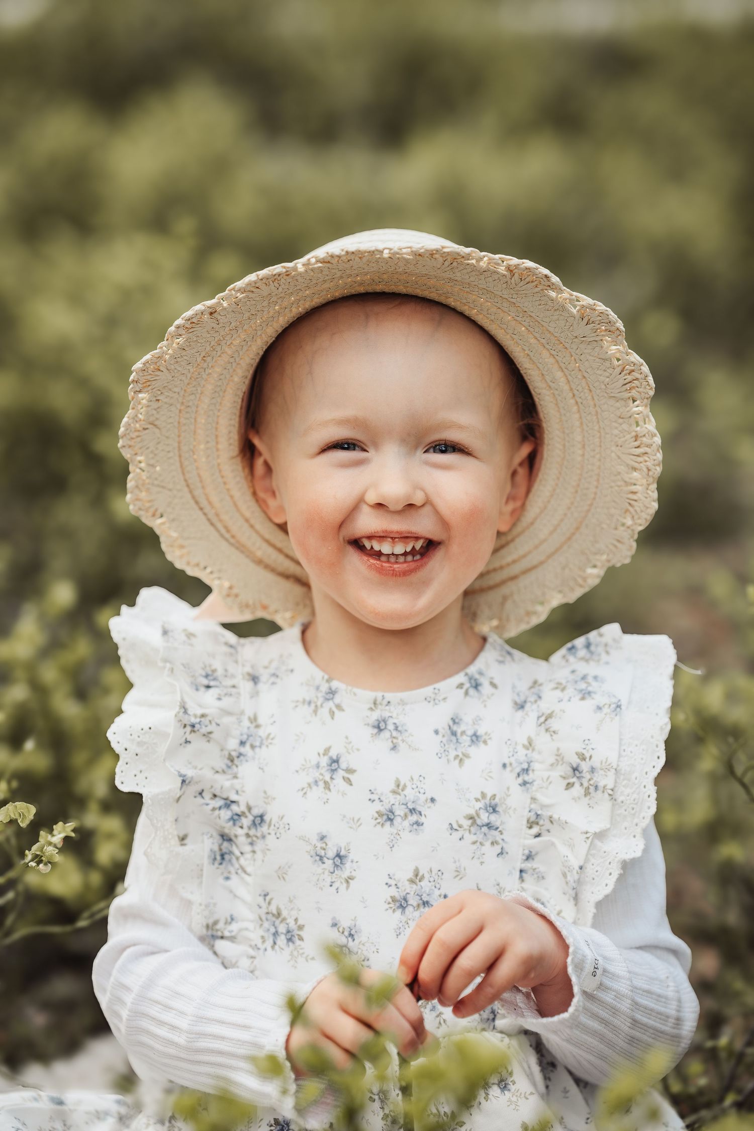 Smiling toddler girl wearing a straw sun hat and floral white dress outdoors in a green garden setting.