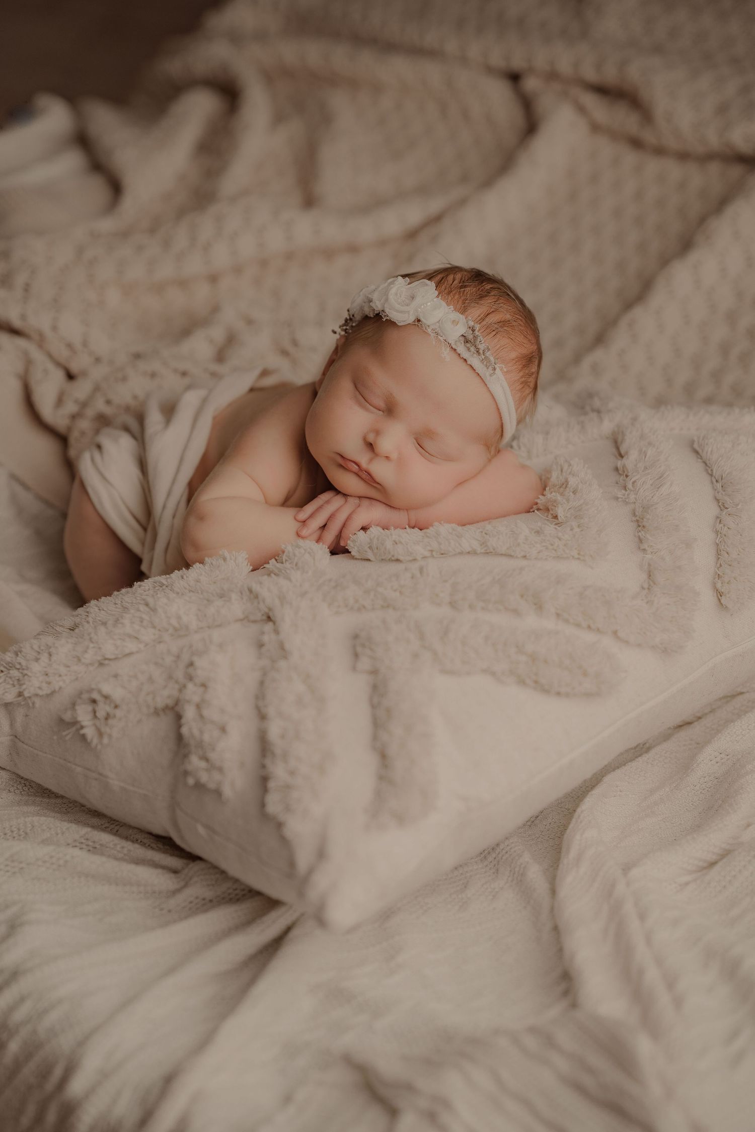 Peaceful newborn baby girl sleeping on soft cream blankets wearing a delicate white floral headband.