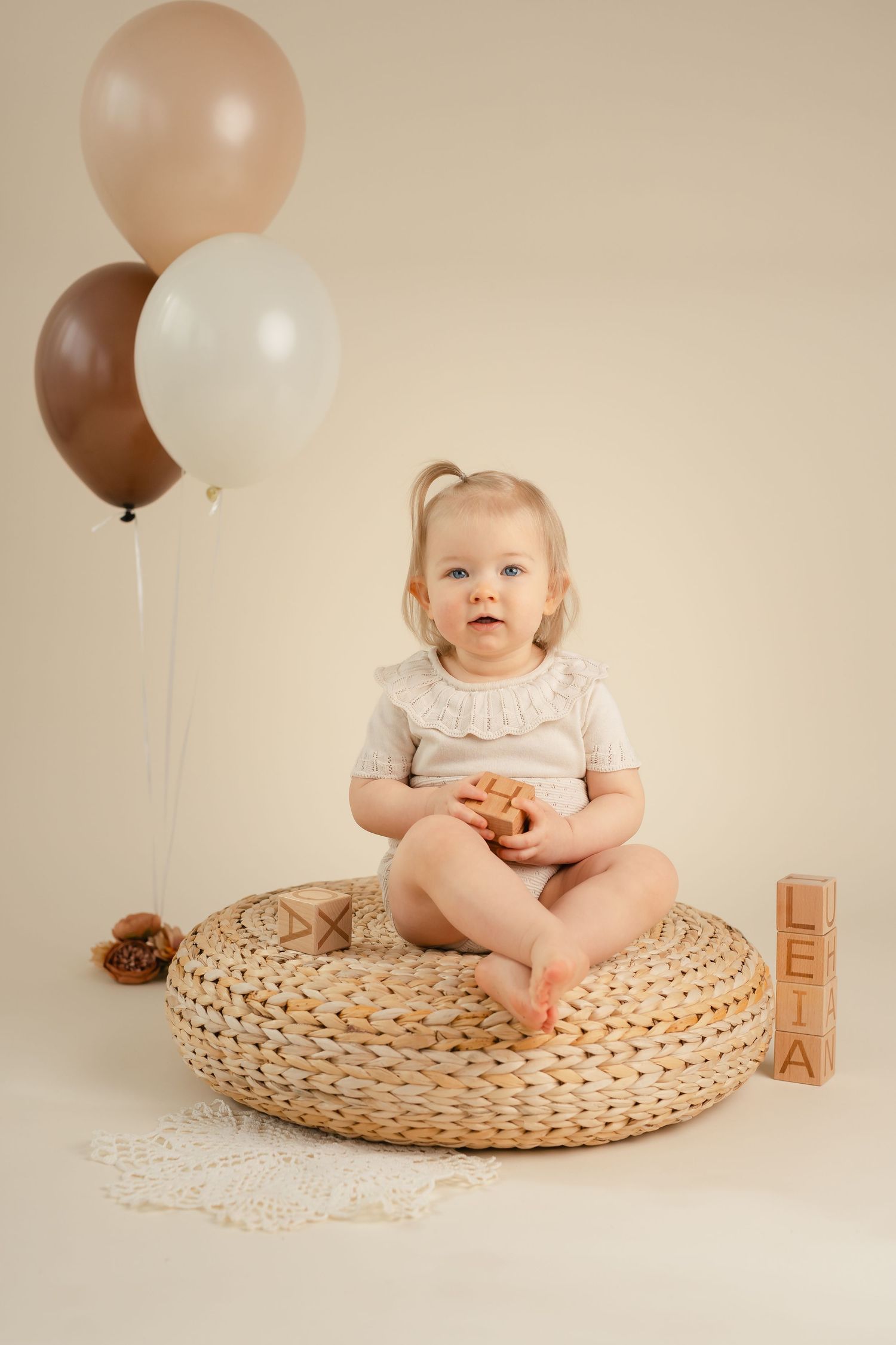 Baby sitting on round woven mat with brown and peach balloons in minimalist first birthday photo session setup.